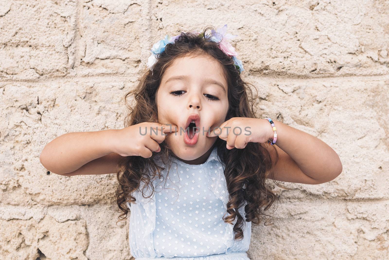 little girl making fun with her face, is in front of a stone wall and wears a blue dress and a flower headband