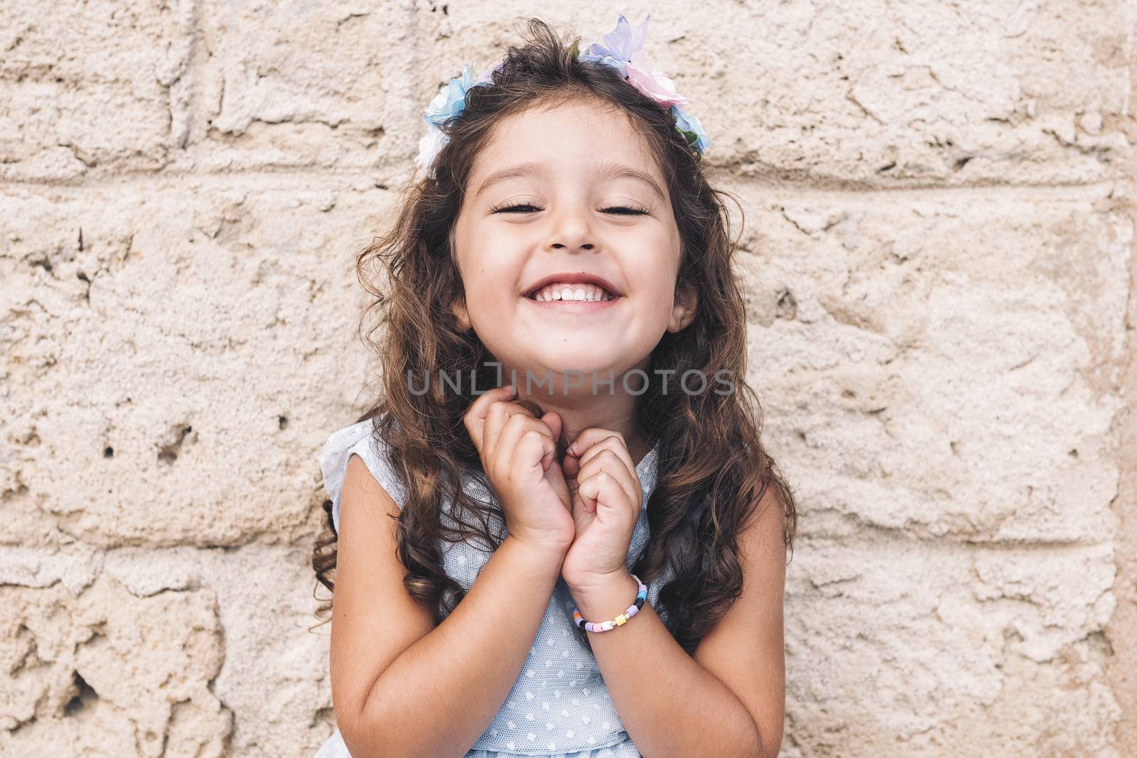 little girl smiles funny, is in front of a stone wall and wears a blue dress and a flower headband