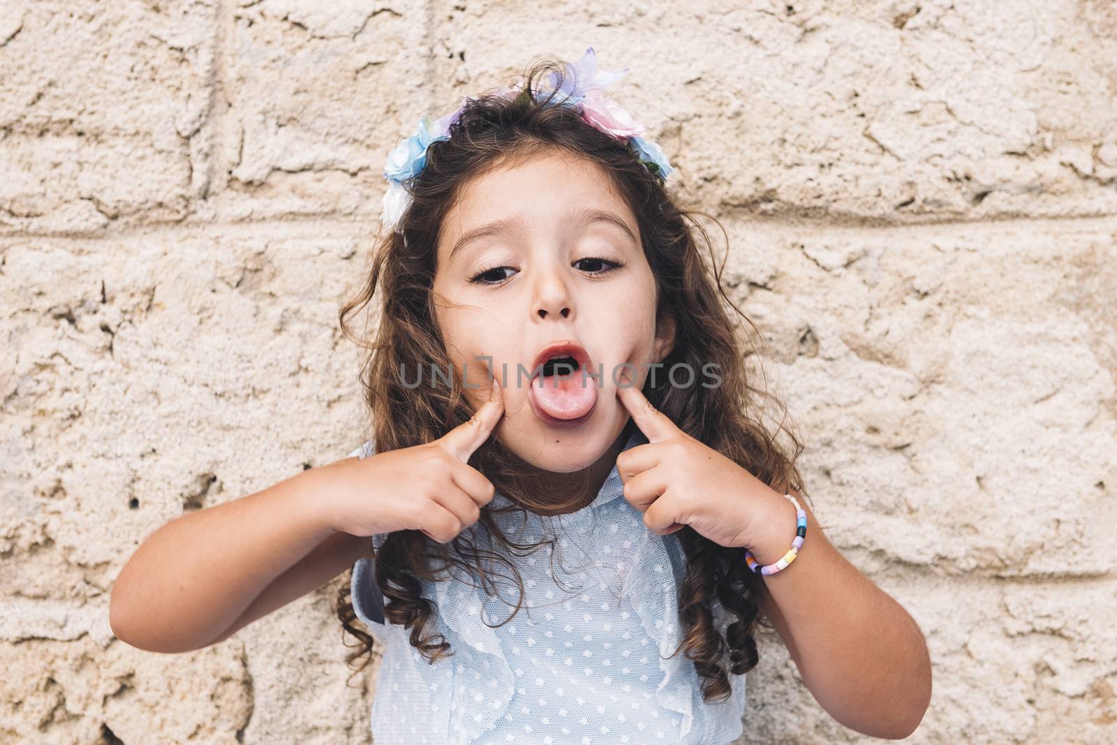 little girl making fun with her tongue, is in front of a stone wall and wears a blue dress and a flower headband