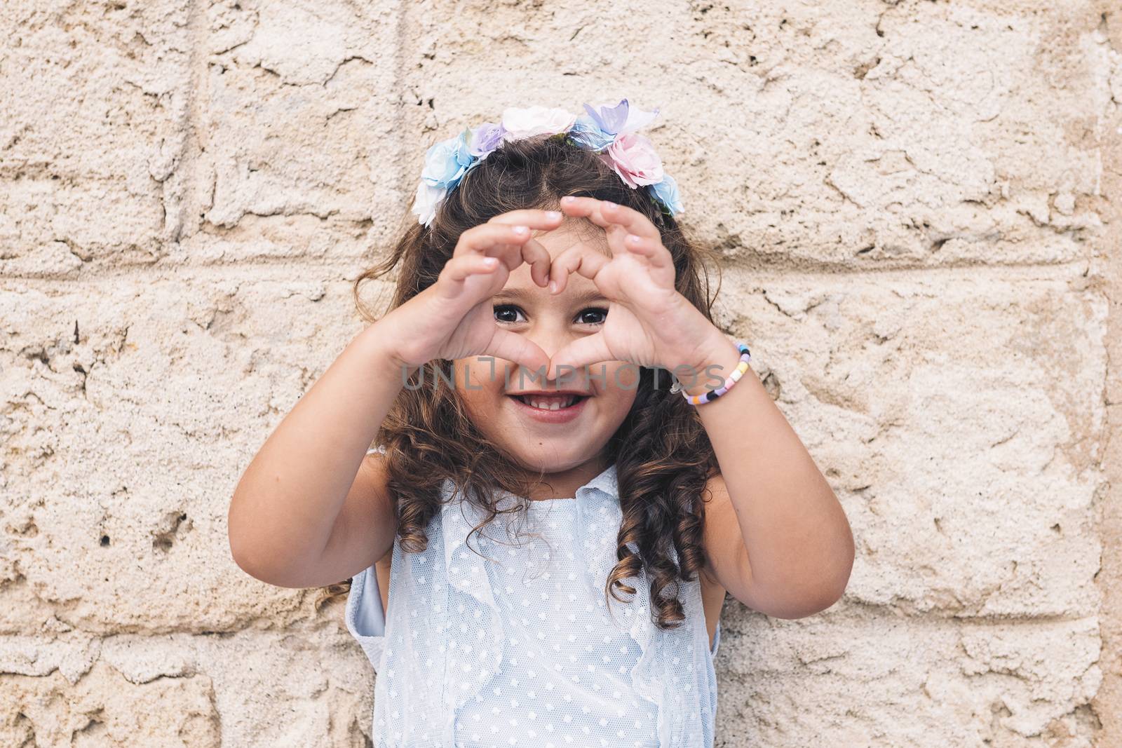 little girl making a heart with her hands, is in front of a stone wall and wears a blue dress and a flower headband