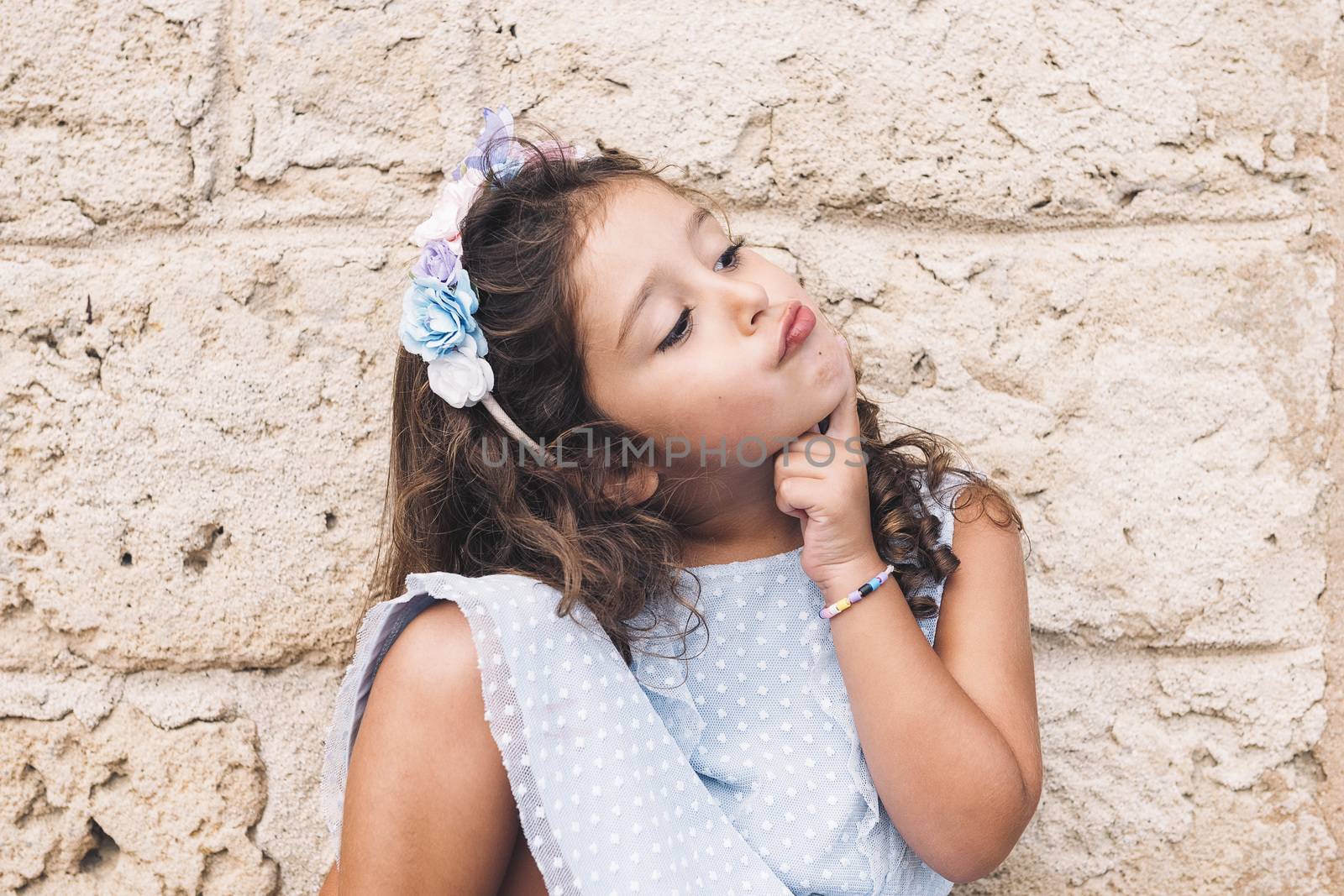 funny little girl doing like she thinks, is in front of a stone wall and wears a blue dress and a flower headband