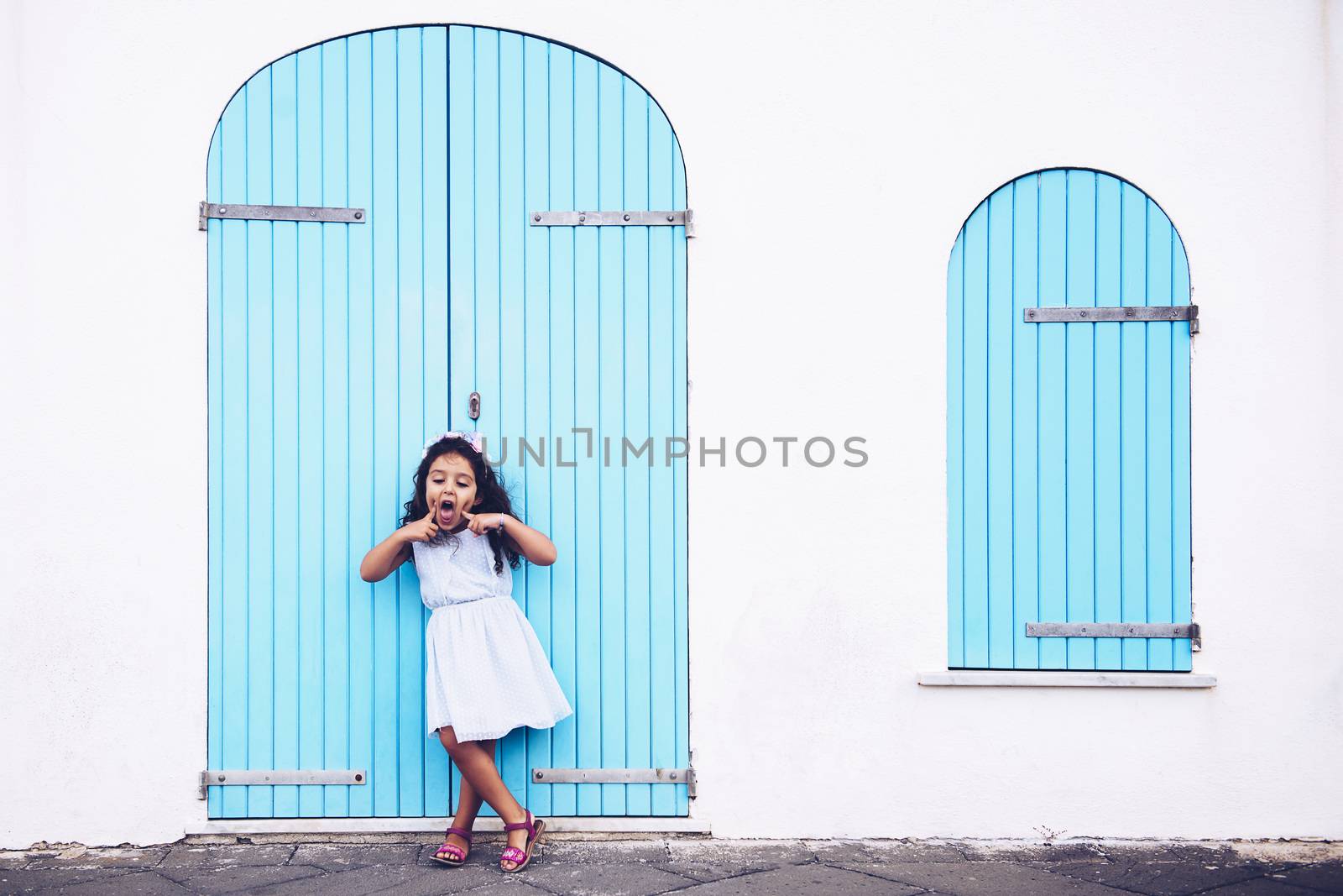 funny little girl making fun with her mouth, she is in front of a white wall with a door and a turquoise wooden window