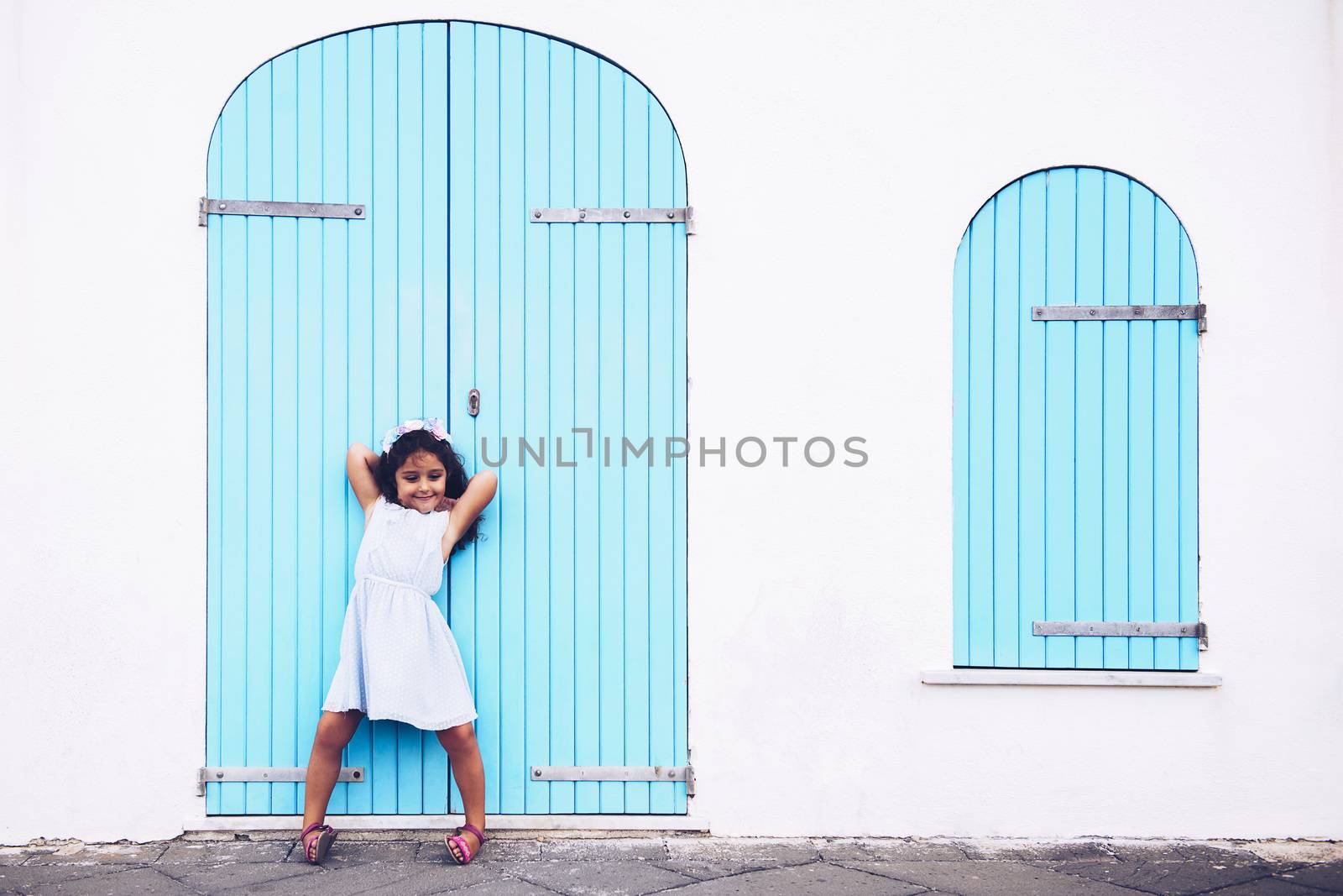 funny little girl making fun, she is in front of a white wall with a door and a turquoise wooden window