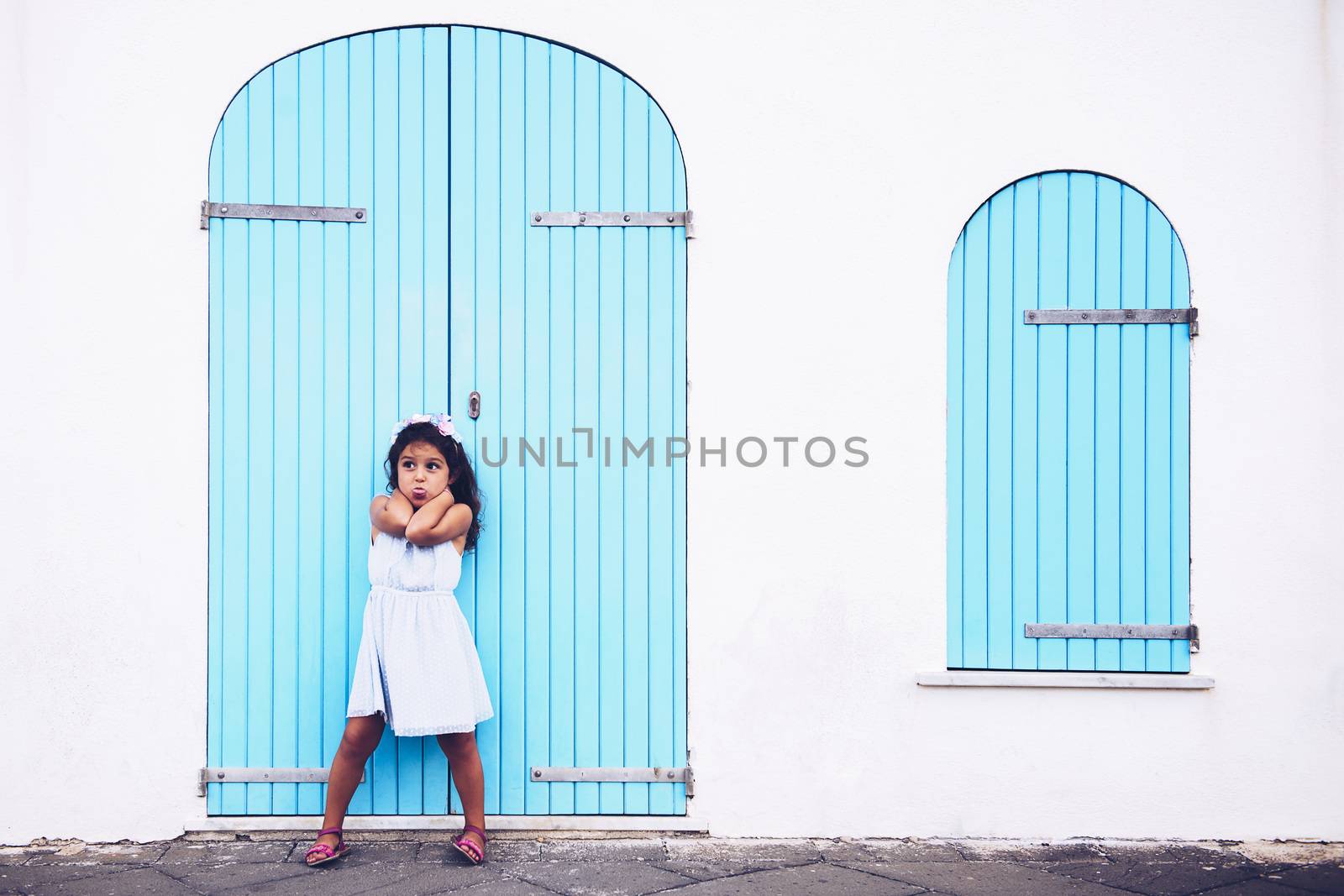 funny little girl making fun by sticking out her tongue, she is in front of a white wall with a door and a turquoise wooden window