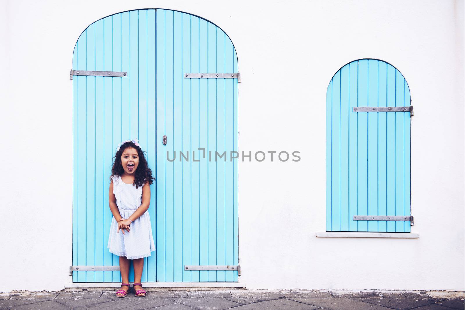 Funny little girl shows tongue, she is in front of a white wall with a door and a turquoise wooden window