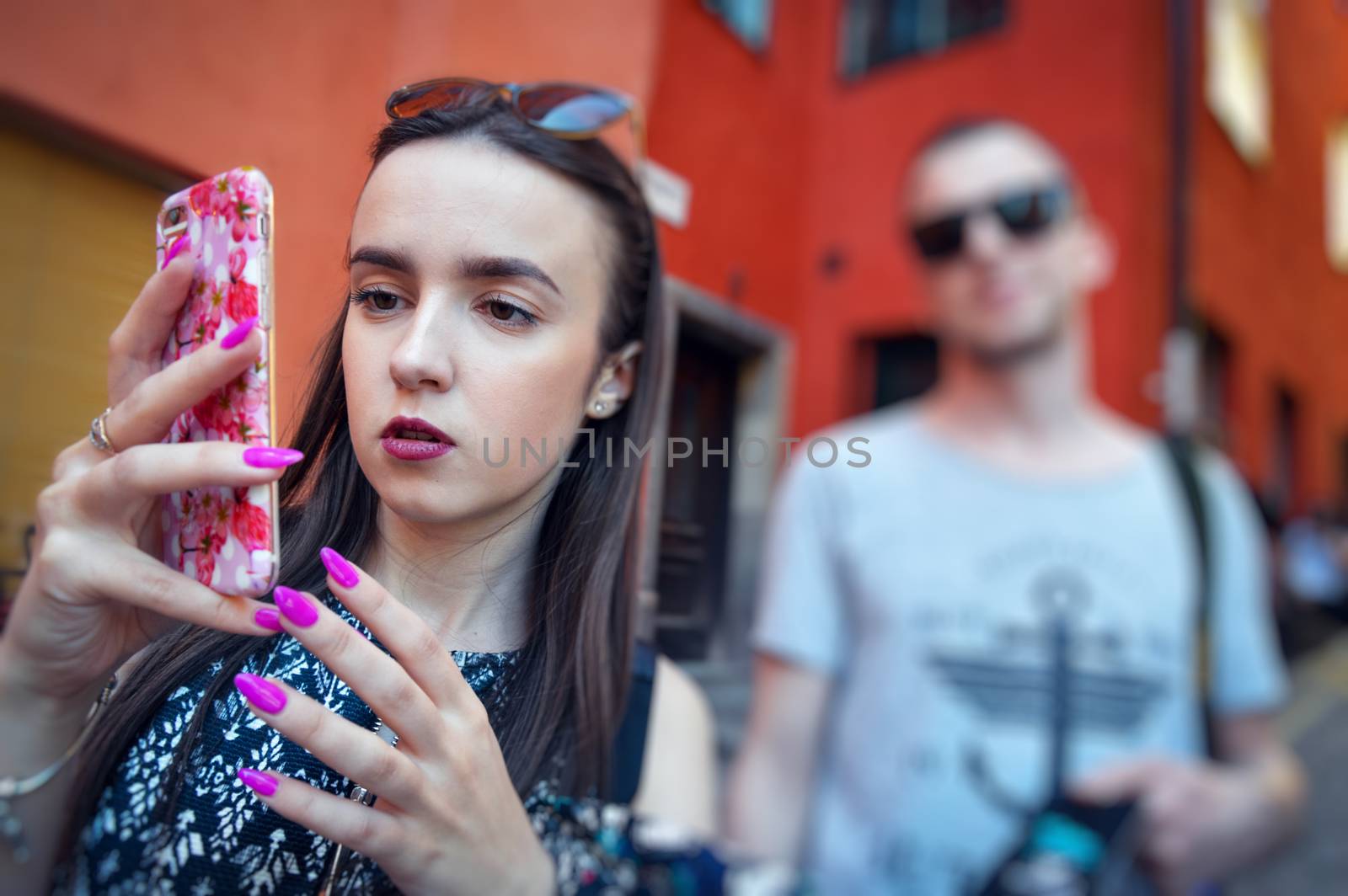 Woman looks into the phone and walks down the street with a man behind. Model wearing stylish sunglasses. City lifestyle. Female fashion concept.