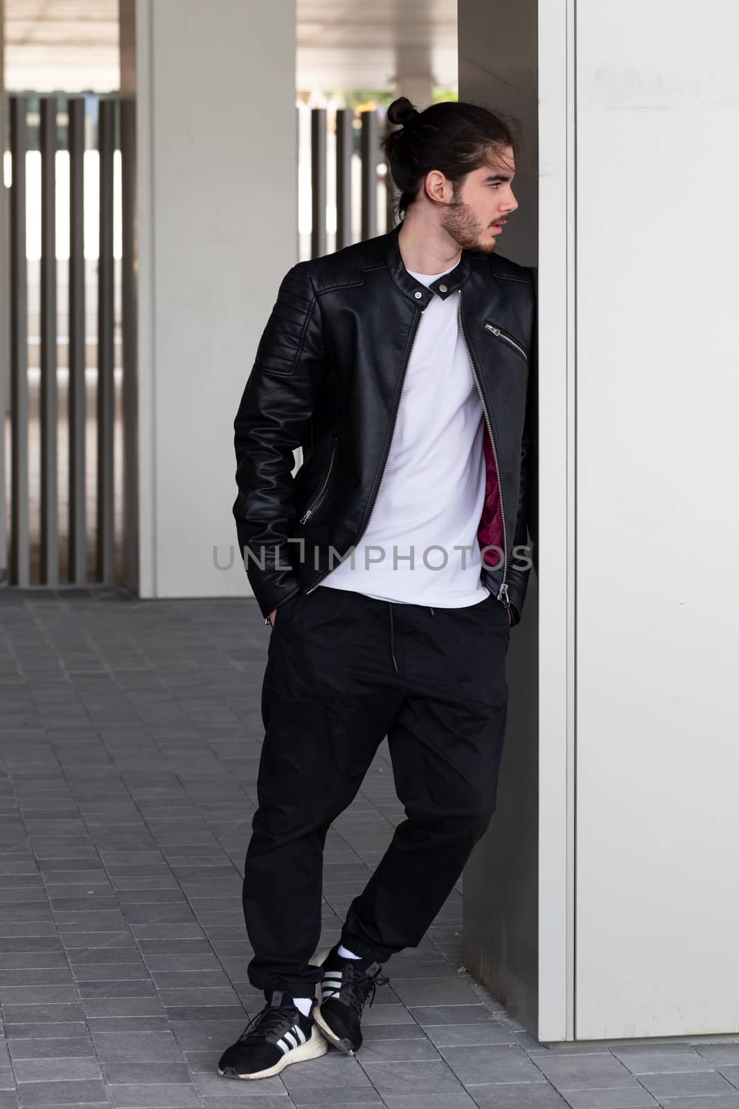 Bearded handsome young man in a black leather jacket with a stylish haircut stands and leans on the wall, looking away.