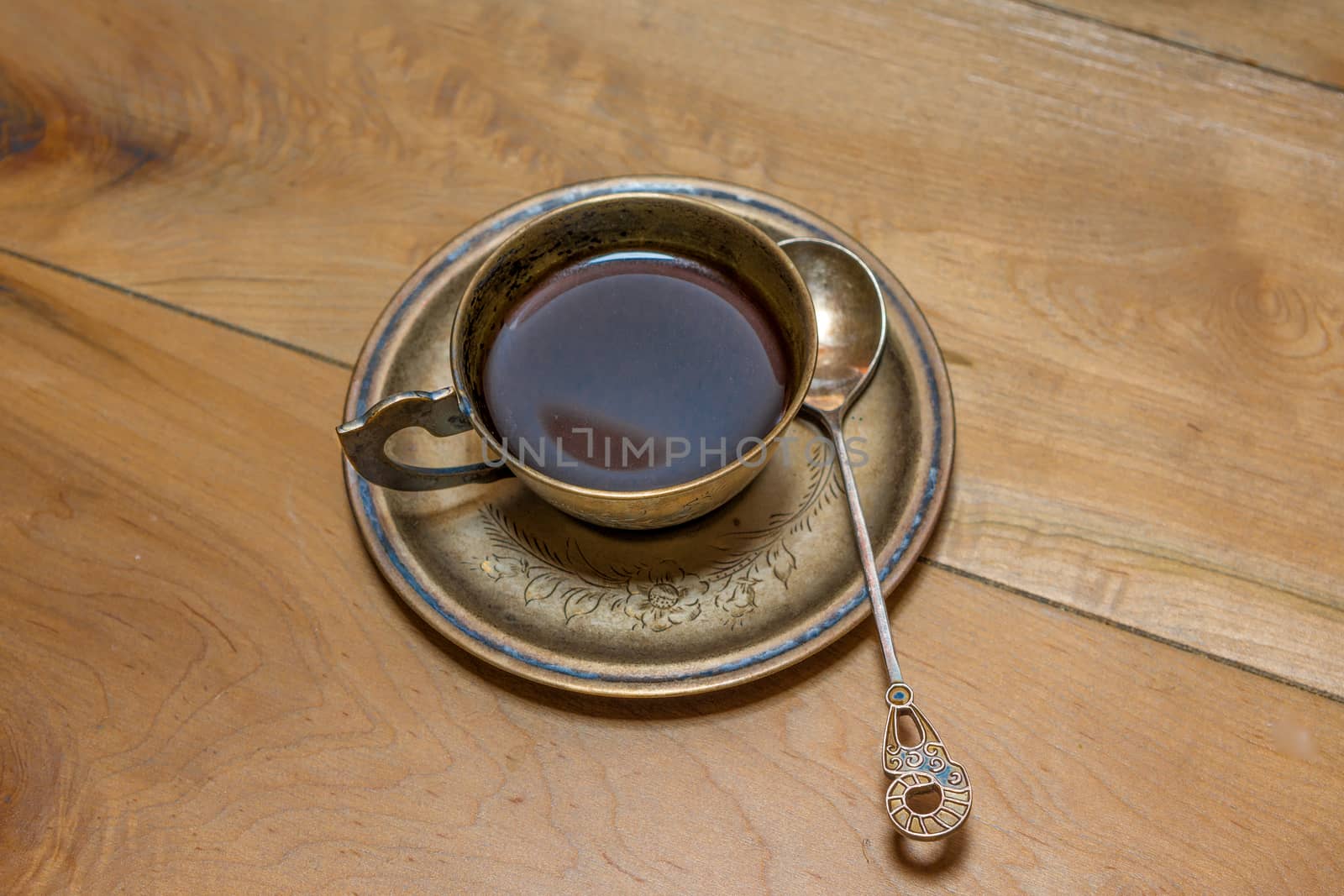 A cup with black coffee, a saucer and a spoon made of bronze stand on an oak table