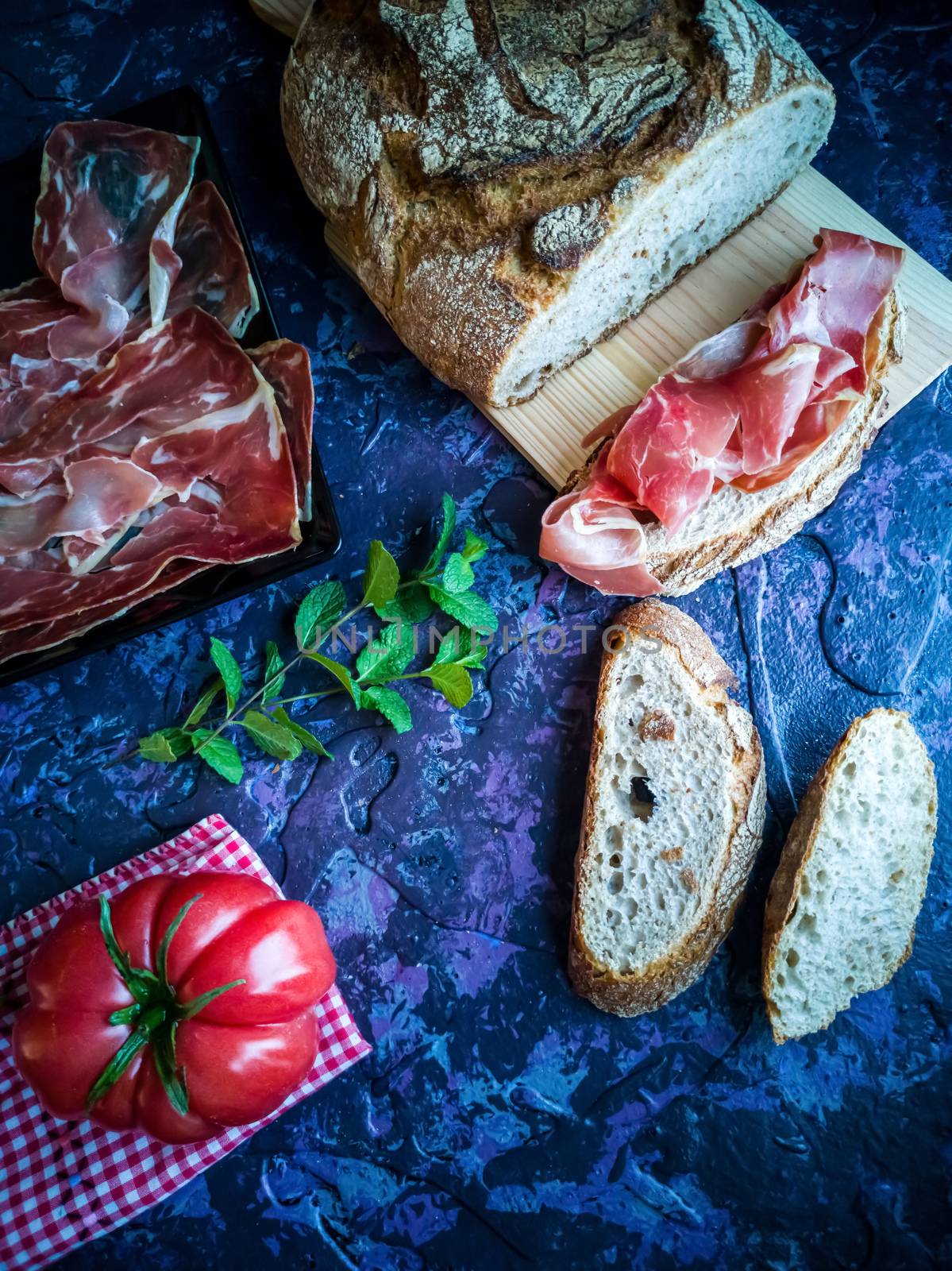 Ham, tomato, bread and herbs in composition on dark background with rough texture
