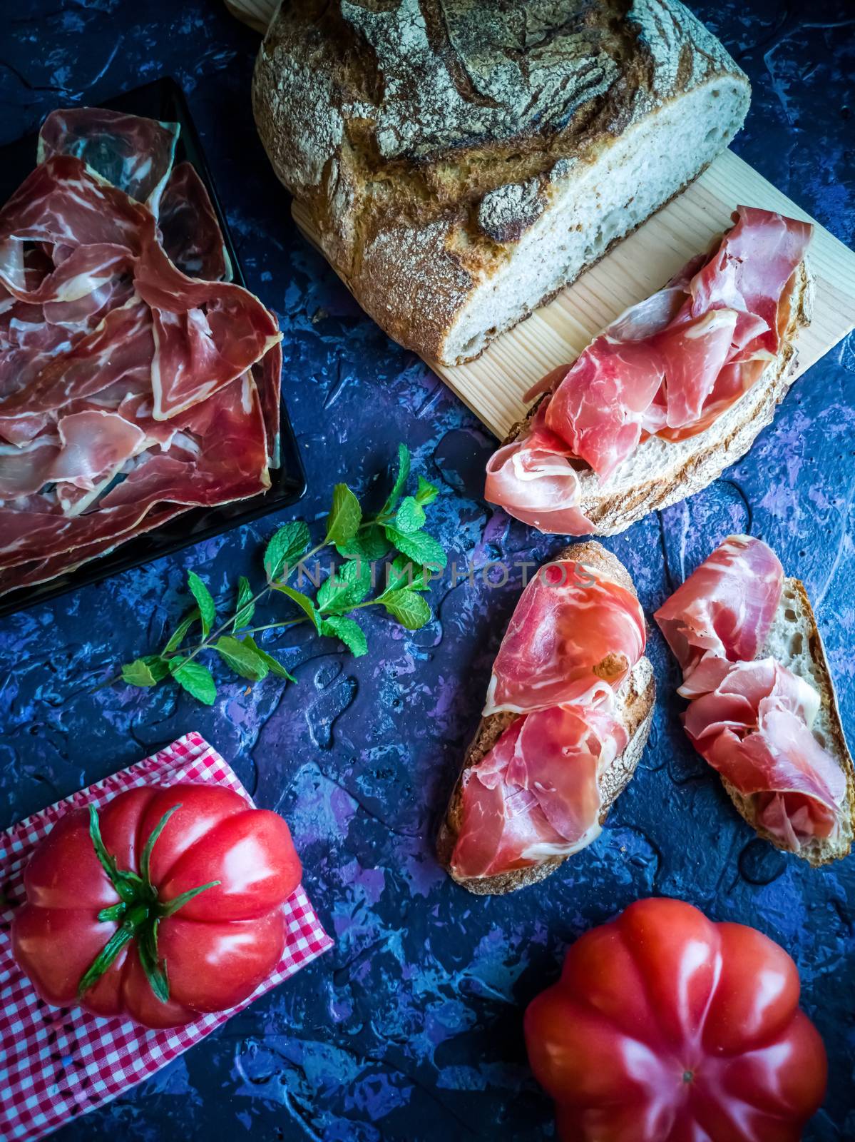Ham, tomato, bread and herbs in composition on dark background with rough texture