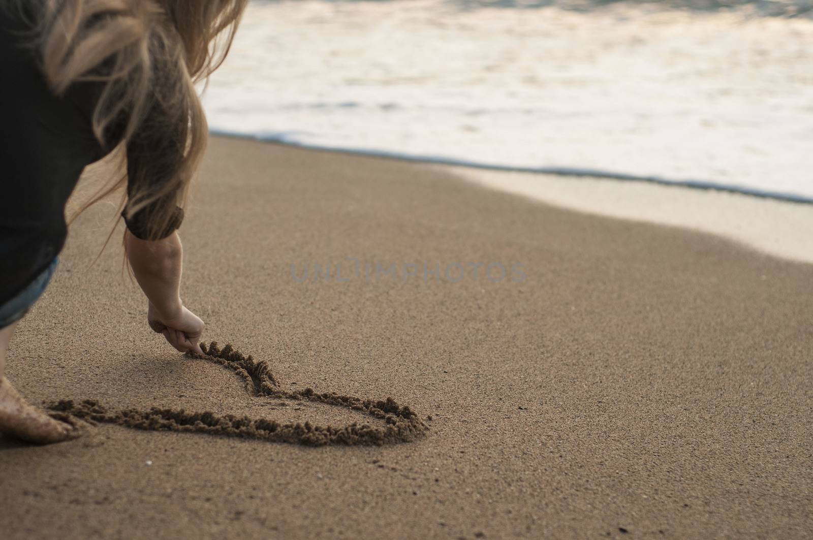 Young girl draws a heart in sand, sitting on beach by claire_lucia