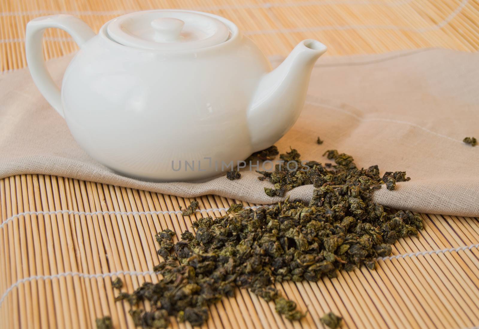 Teapot with green tea leaves on the napkin, bamboo background.