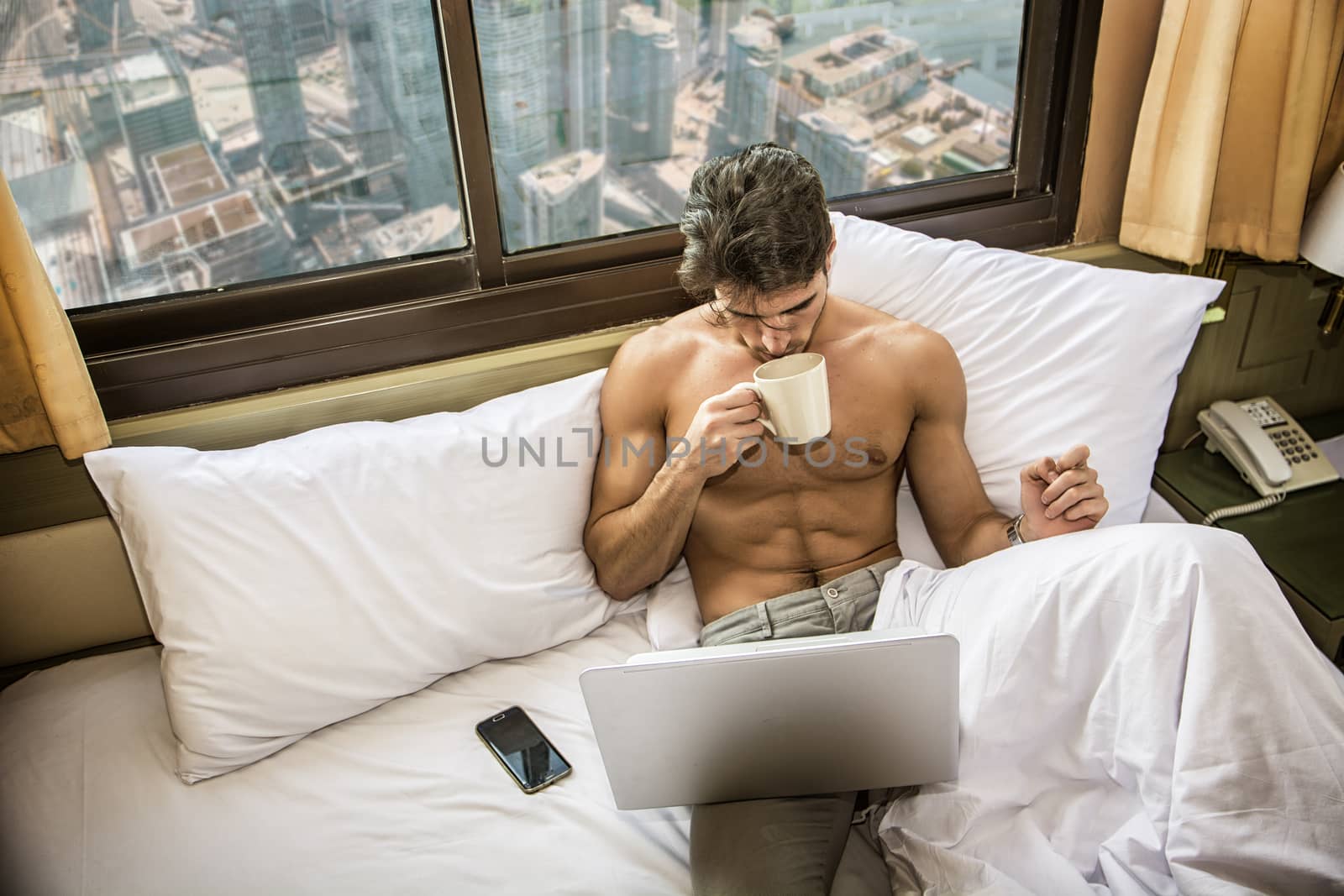 Naked young man with muscular body on bed with mug or cup in hand with coffee or tea, looking at laptop computer screen