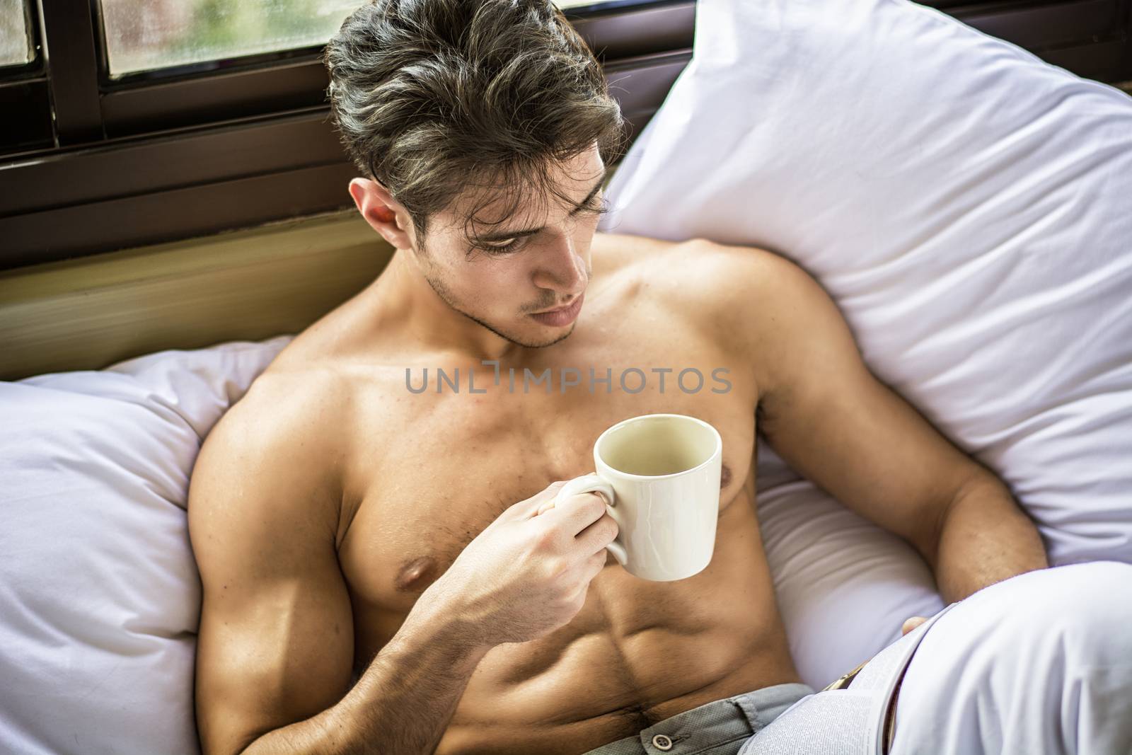 Sexy handsome young man laying shirtless on his bed next to window, holding a coffee or tea cup while reading a book