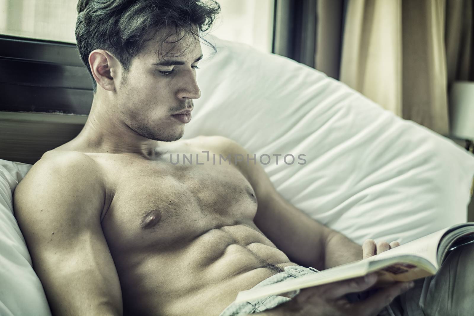 Sexy handsome young man laying shirtless on his bed next to window, reading a book