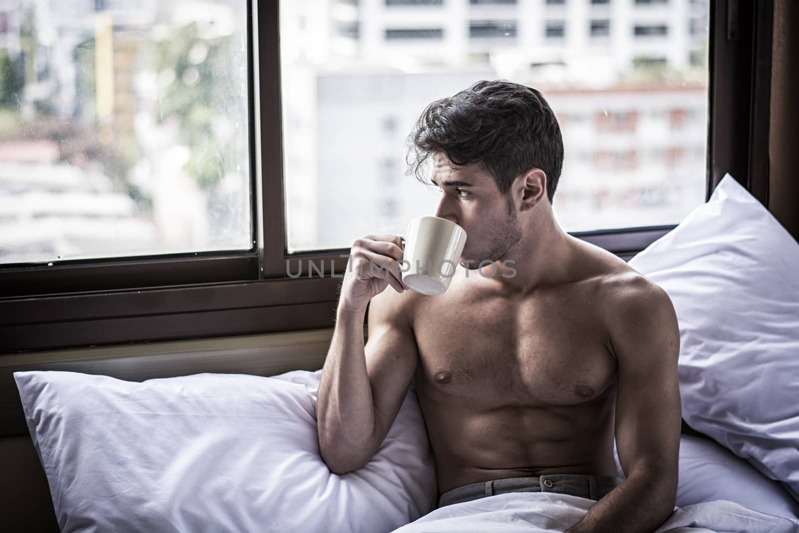 Naked young man with muscular body on bed with mug or cup in hand with coffee or tea