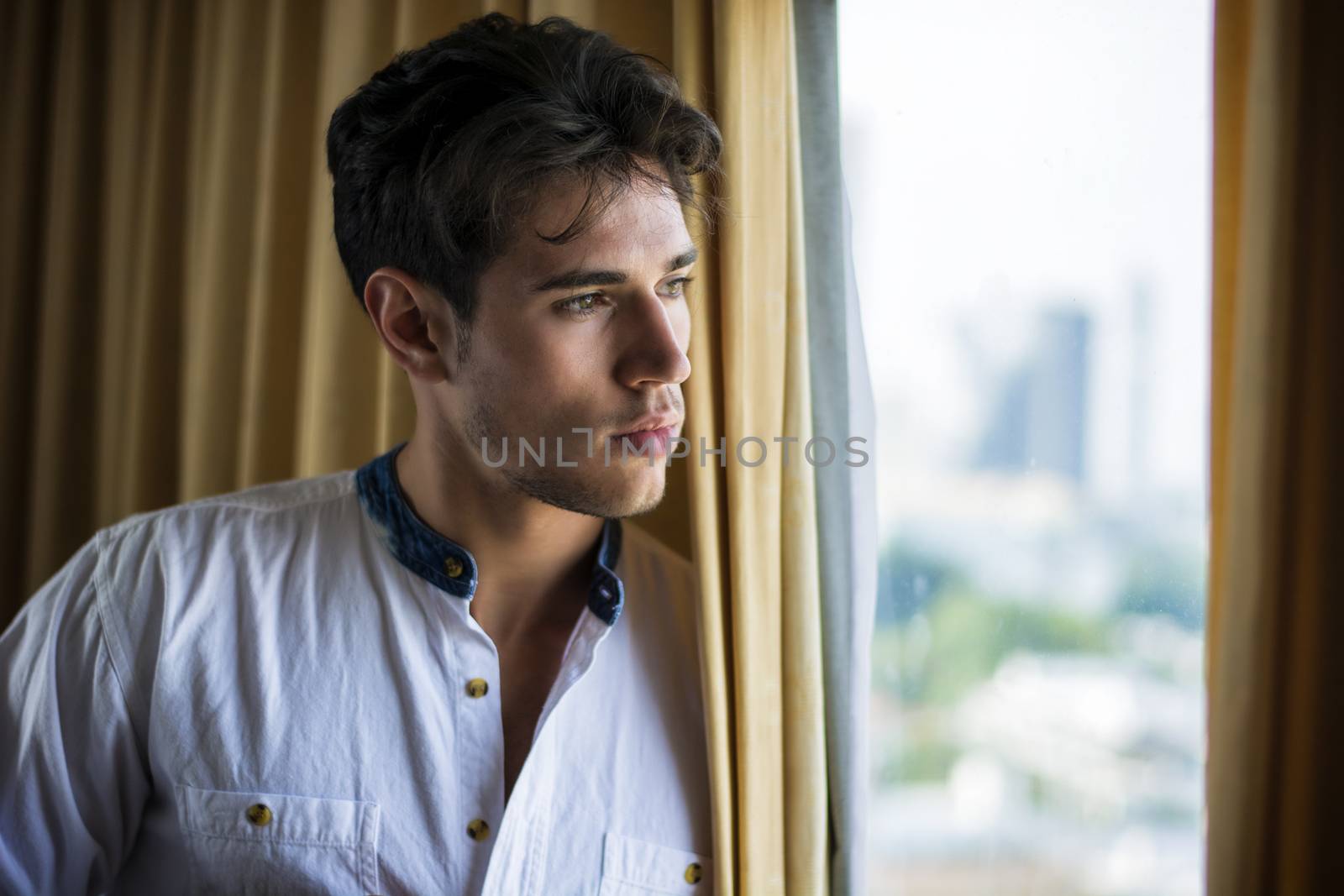 Sexy handsome young man standing and dressing, in his bedroom next to window curtains