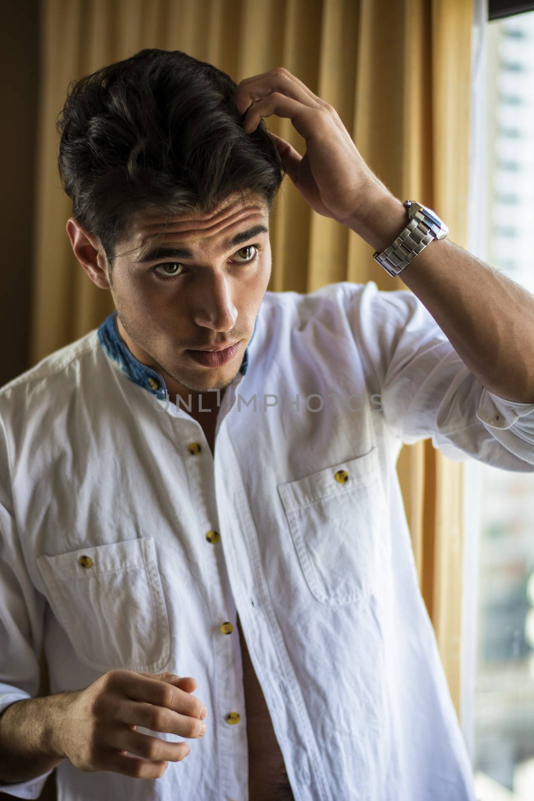 Sexy handsome young man standing and dressing, in his bedroom next to window curtains
