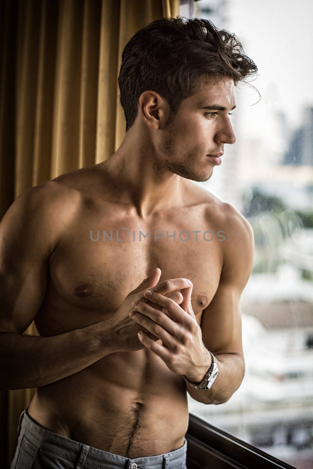 Sexy handsome young man standing shirtless in his bedroom next to window curtains