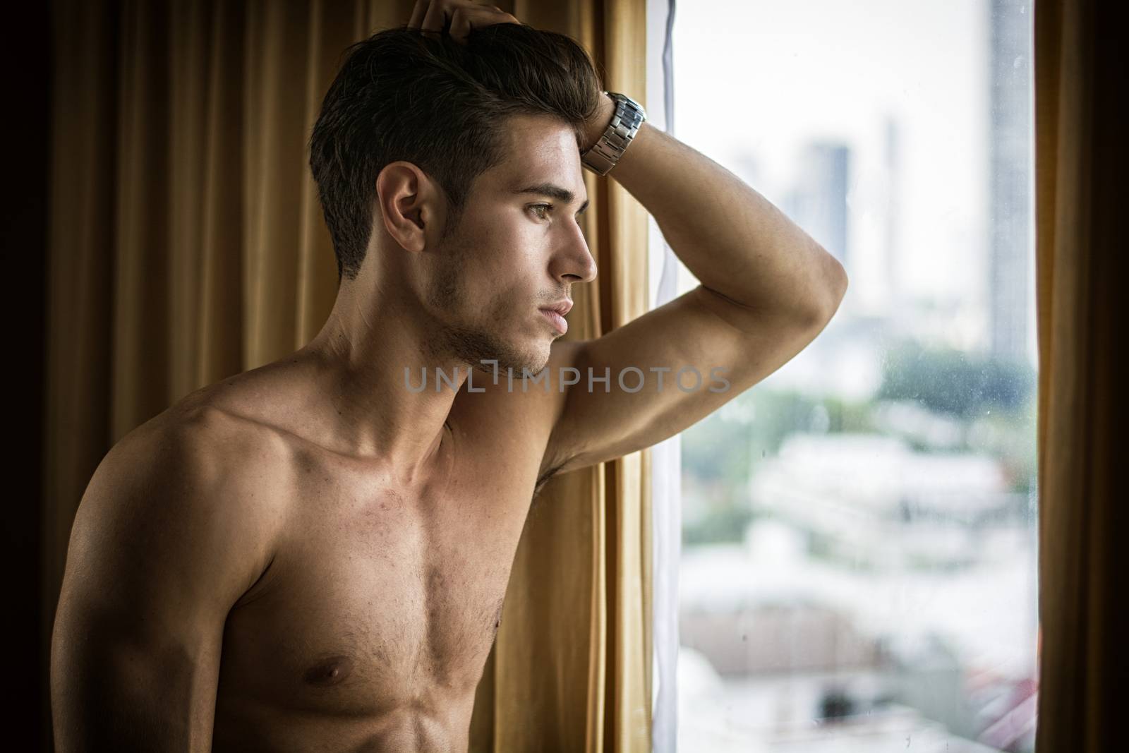 Sexy handsome young man standing shirtless in his bedroom next to window curtains