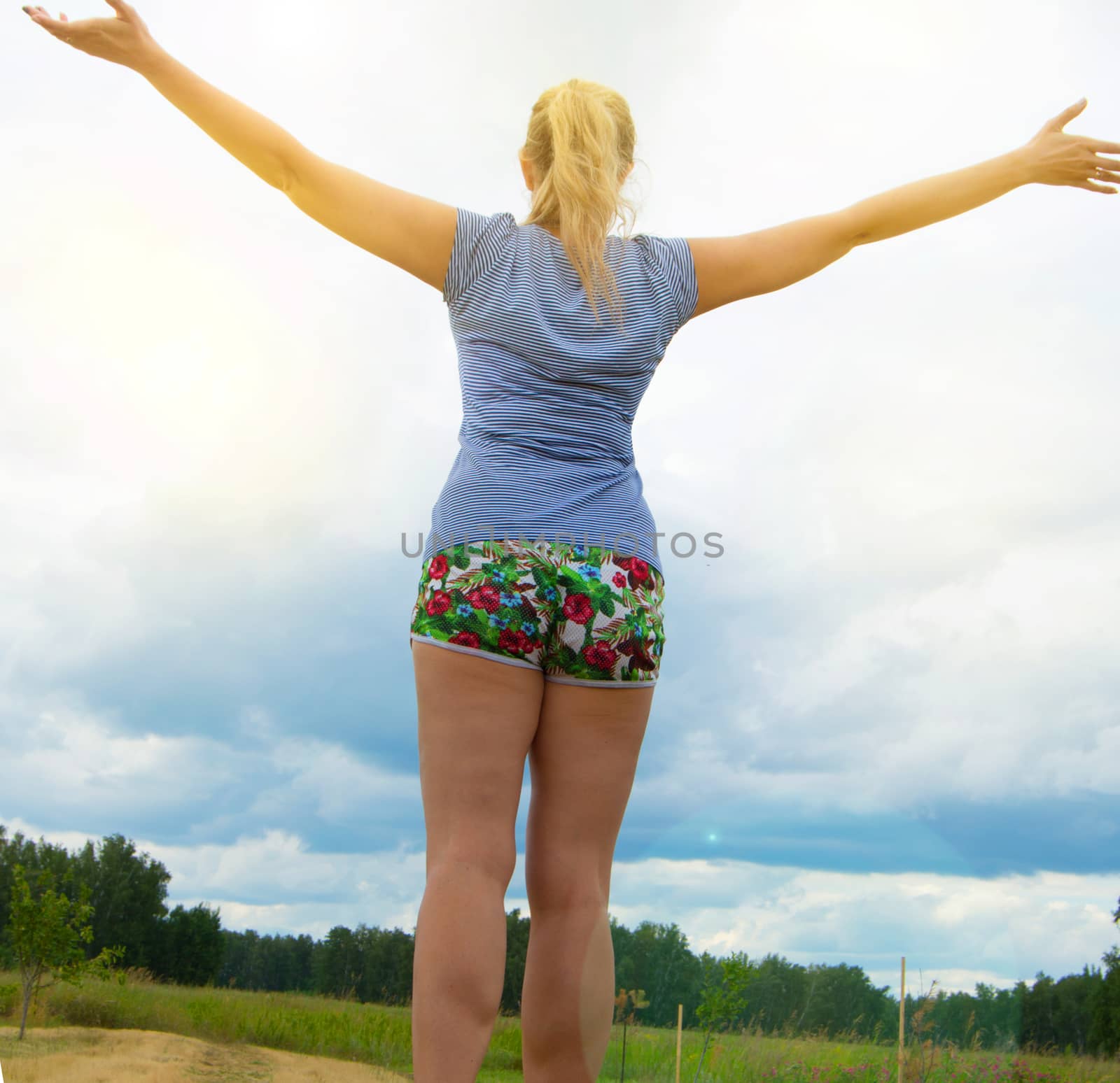 Happy young blonde wearing shorts and a t-shirt stands on the road against the sky with her arms outstretched, illuminated by sunlight on a summer day by claire_lucia