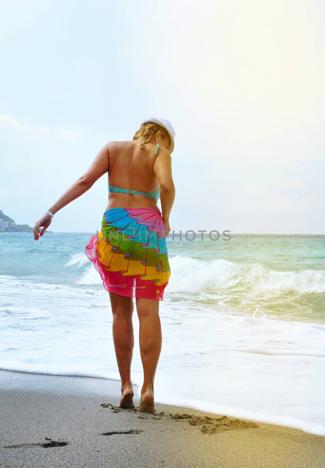 Attractive sexy young woman walks on the beach in a bikini and colorful pareo, luxurious summer vacation, vertical shot, sun rays and glare by claire_lucia