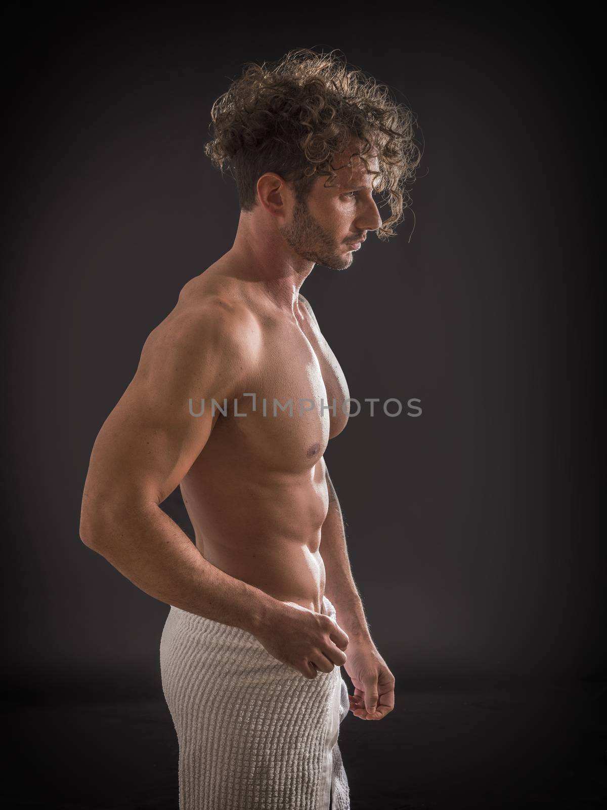 Portrait of naked handsome young man with languishing look covering crotch with a towel, against black background