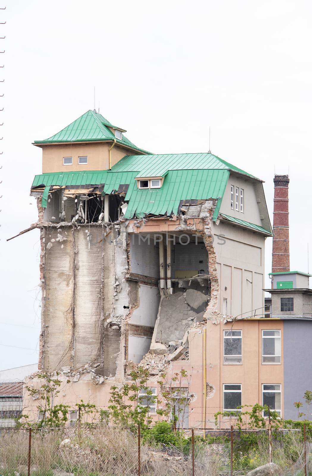 View on a partially collapsed brick industrial building with green metal roof and light sky above. Industrial silo building.