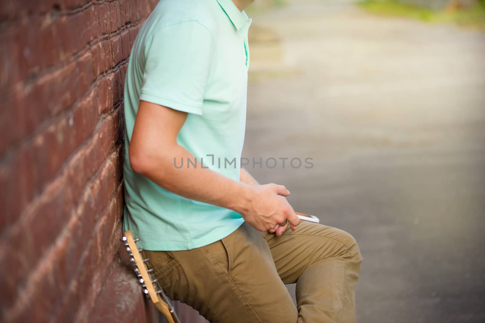 Summer, party, young stylish guy standing near brown brick wall, hands holding mobile phone next to guitar, sunlight.