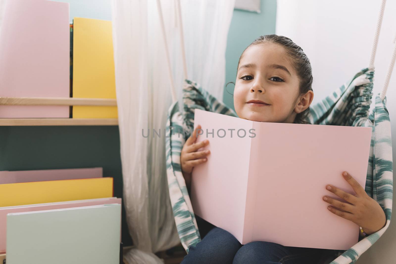 little girl is sitting on the hammock and reading a book near the window, funny lovely child having fun in kids room