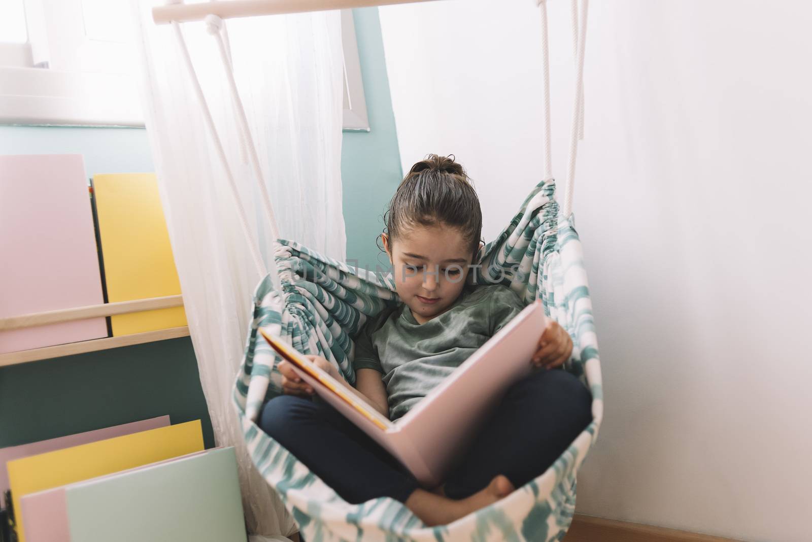relaxed little girl reading a book in her hammock near the window, funny lovely child having fun in kids room, copy space for text
