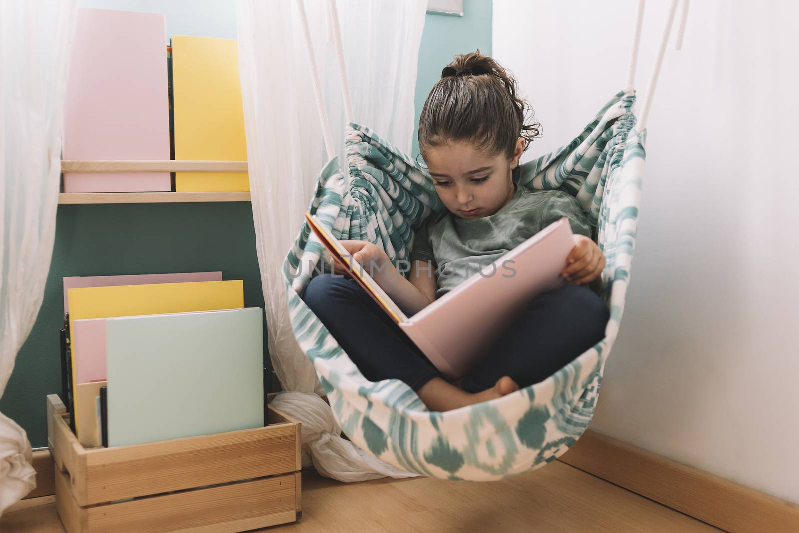 relaxed little girl reading a book in her hammock near the window, funny lovely child having fun in her kids room