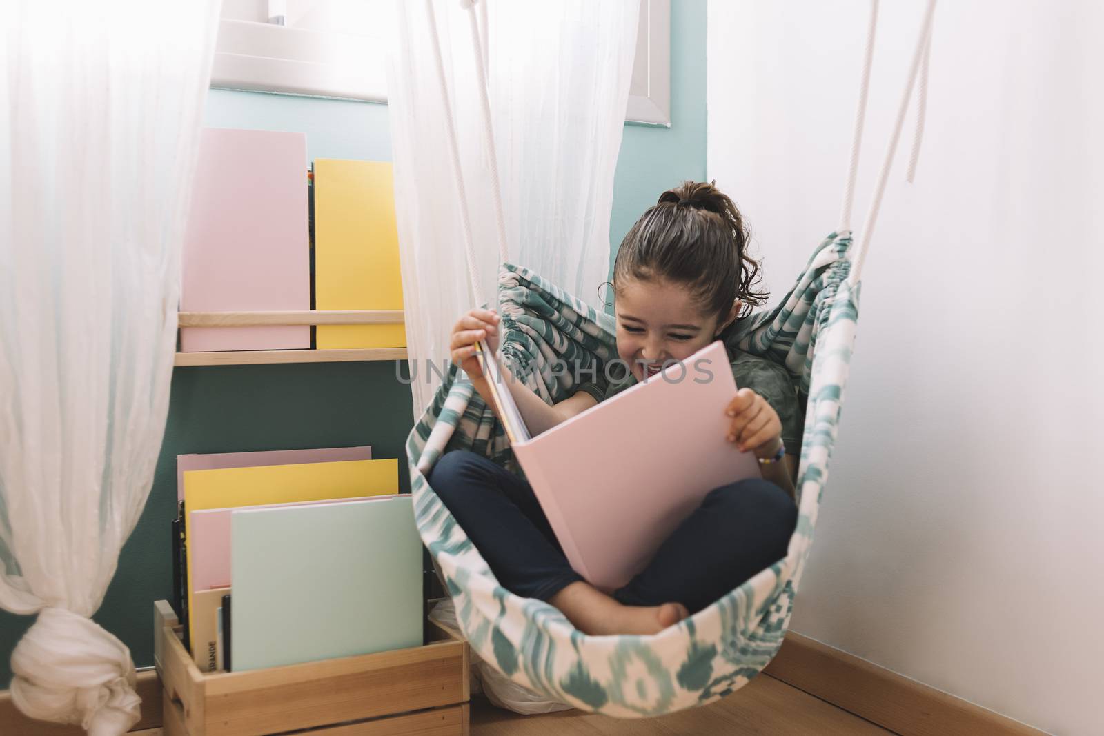 little girl laughing while reading a book in her hammock near the window, funny lovely child having fun in her kids room