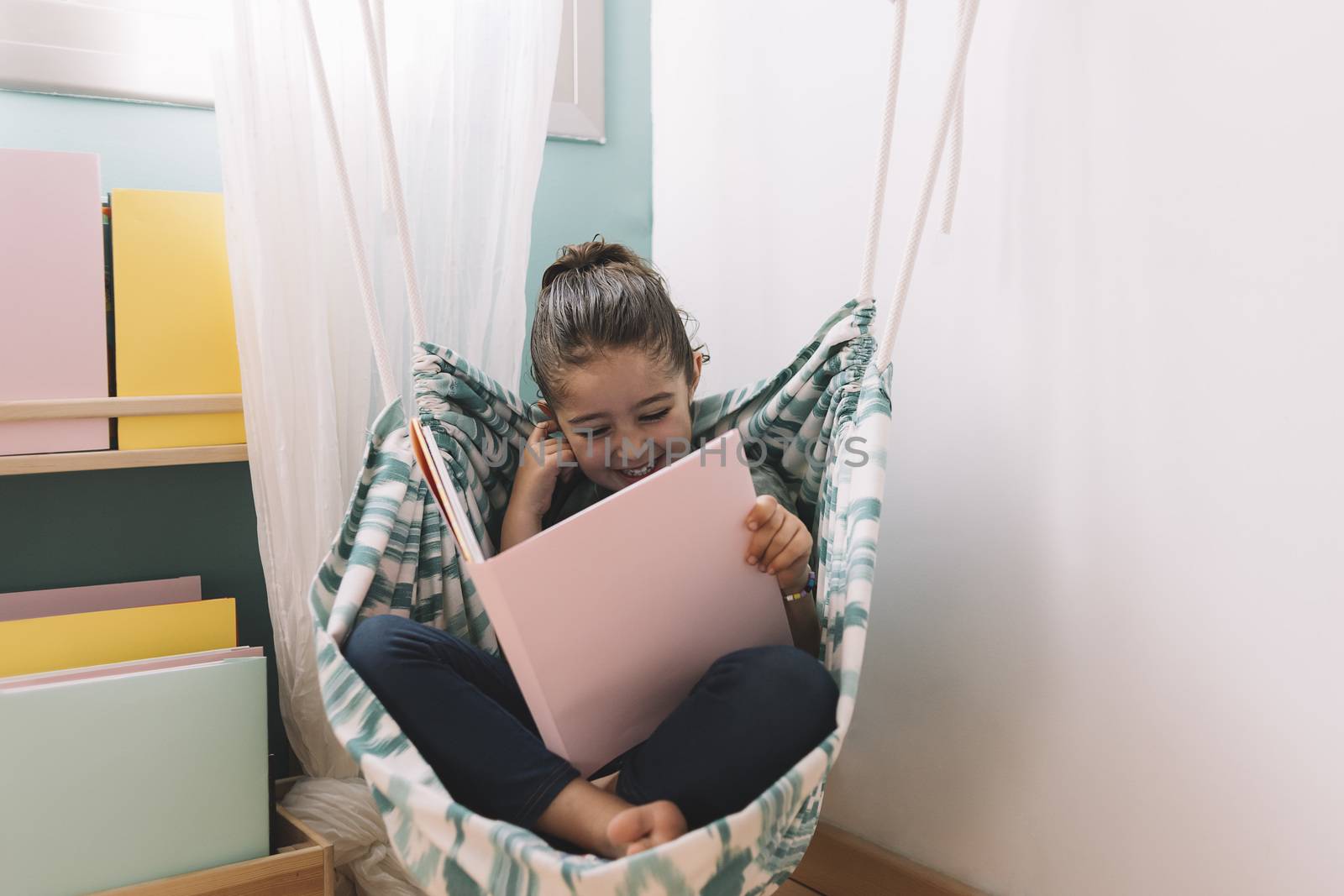 little girl laughing while reading a book near the window, funny lovely child having fun in her kids room, copy space for text