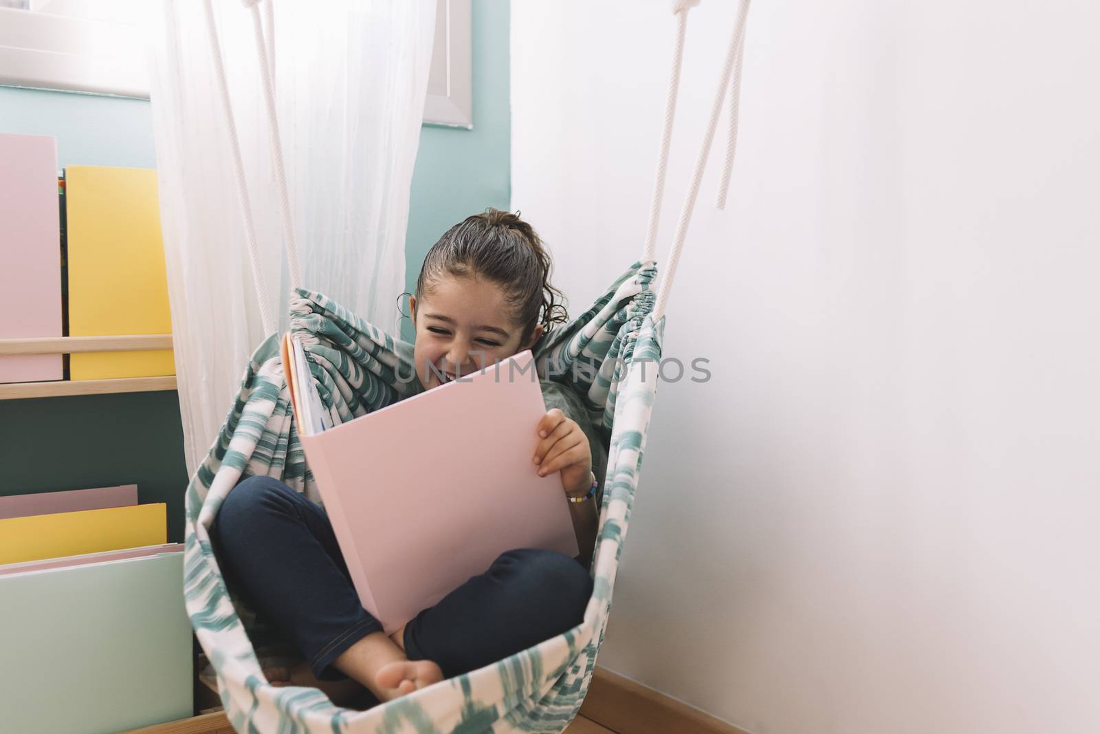 little girl laughing while reading a book in her kids room near the window, funny lovely child having fun at home, copy space for text