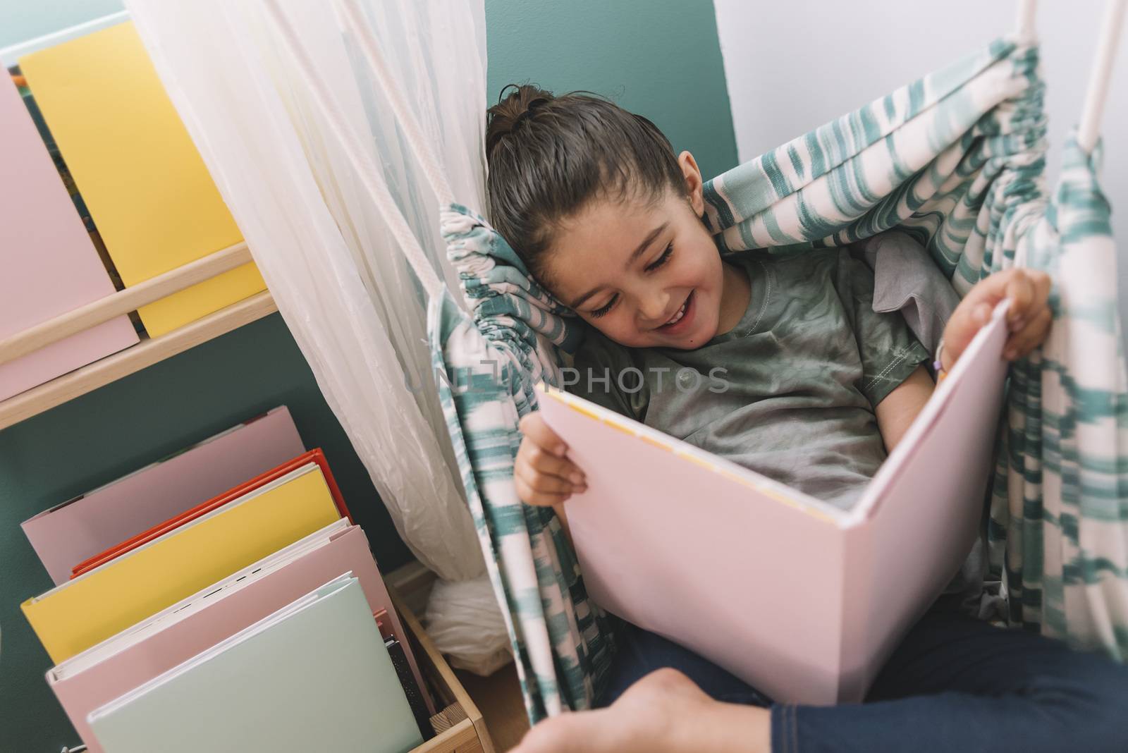 little girl smiling while reading a book in her kids room near the window, funny lovely child having fun at home