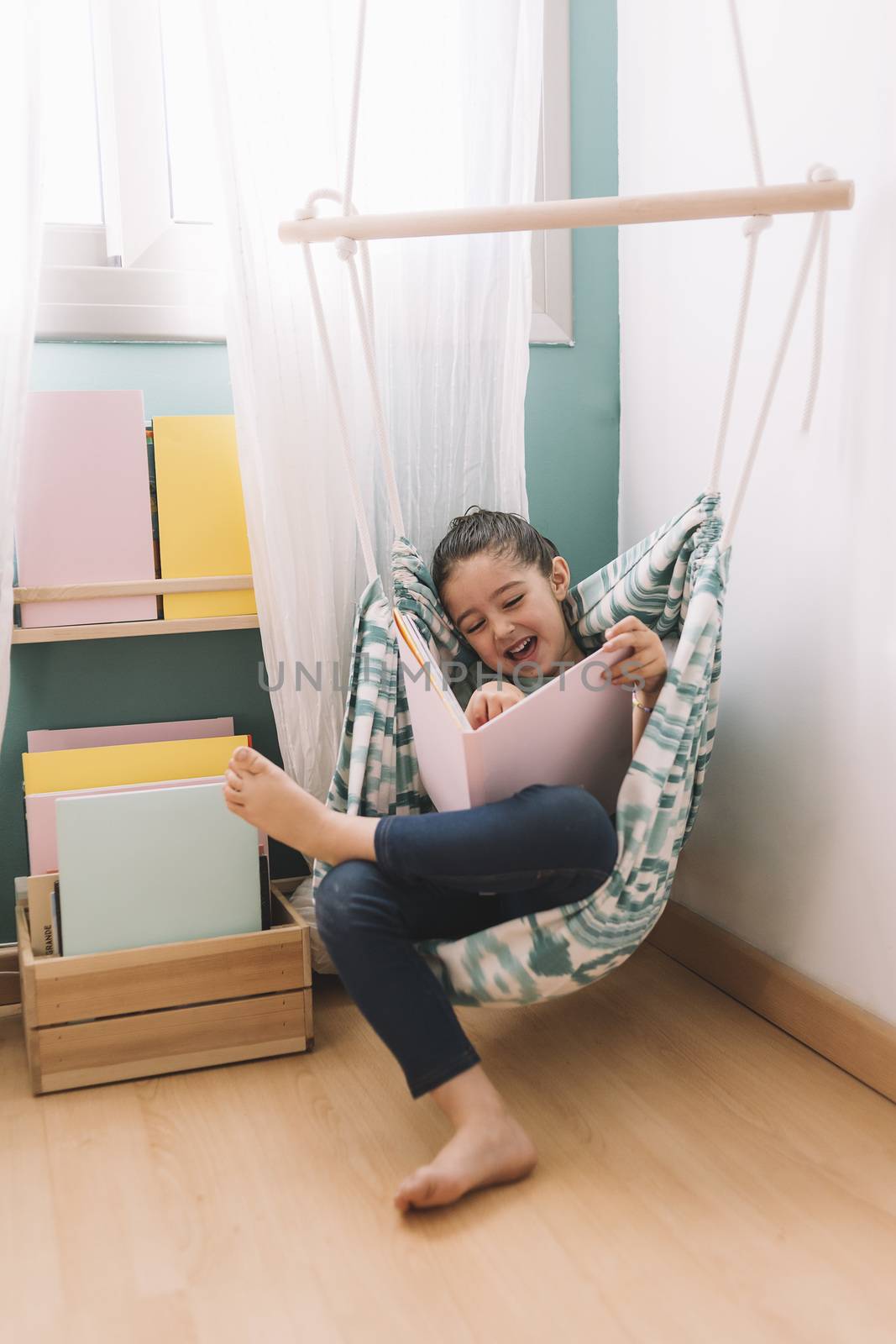 vertical photo of a little girl laughing while reading a book at home near the window, funny lovely child having fun in her kids room, copy space for text