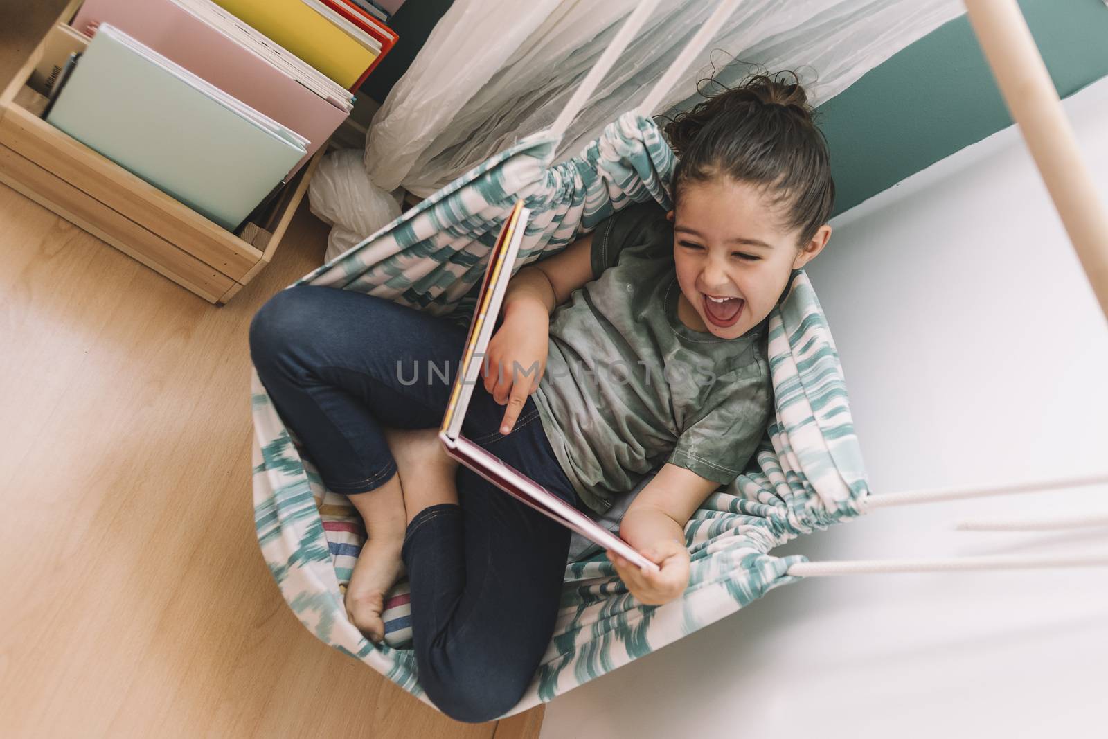 little girl laughs out loud reading a book at home near the window, funny lovely child having fun in her kids room, copy space for text