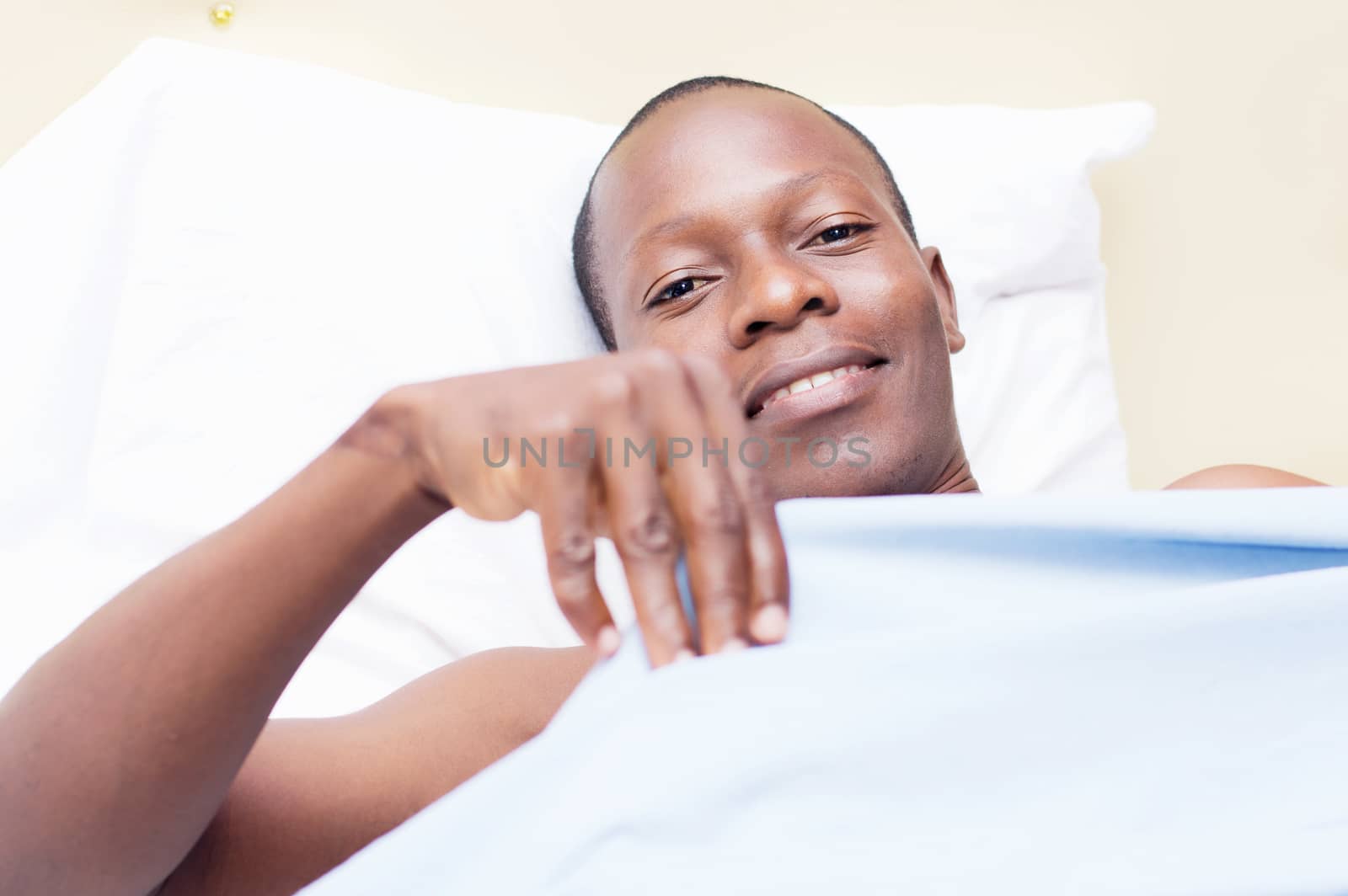 young man lying on a bed, ready to cover smile at the camera.
