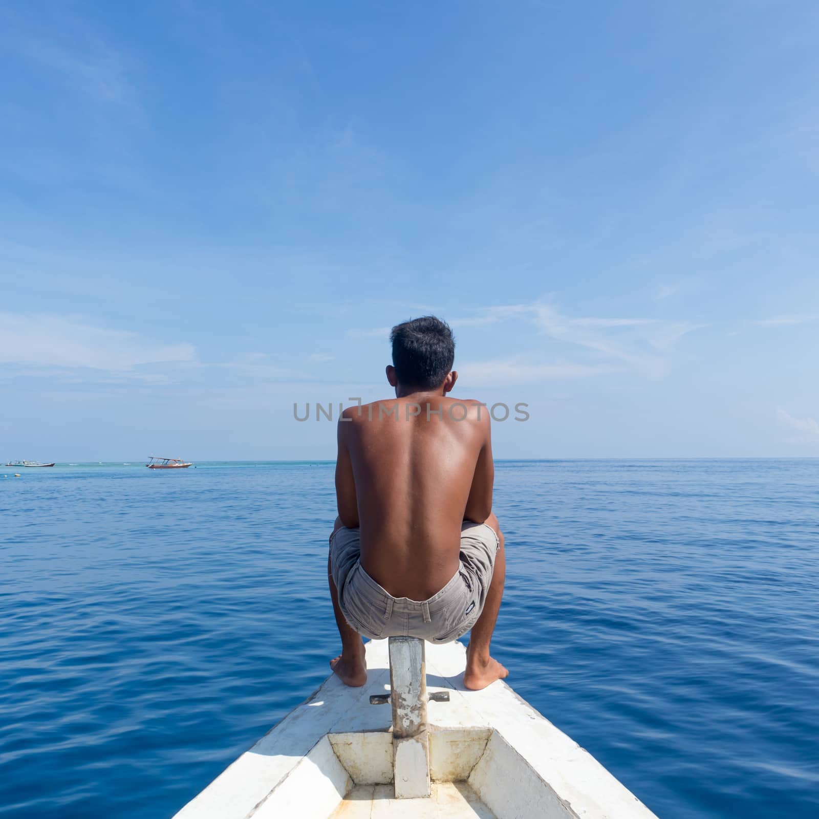 Unrecognizable Local Sporty Guy Sitting Topless at the Bow of Traditional White Wooden Sail Boat, Enjoying the Breeze, Looking At Beautiful Blue Sea of Gili Islands near Bali, Indonesia.