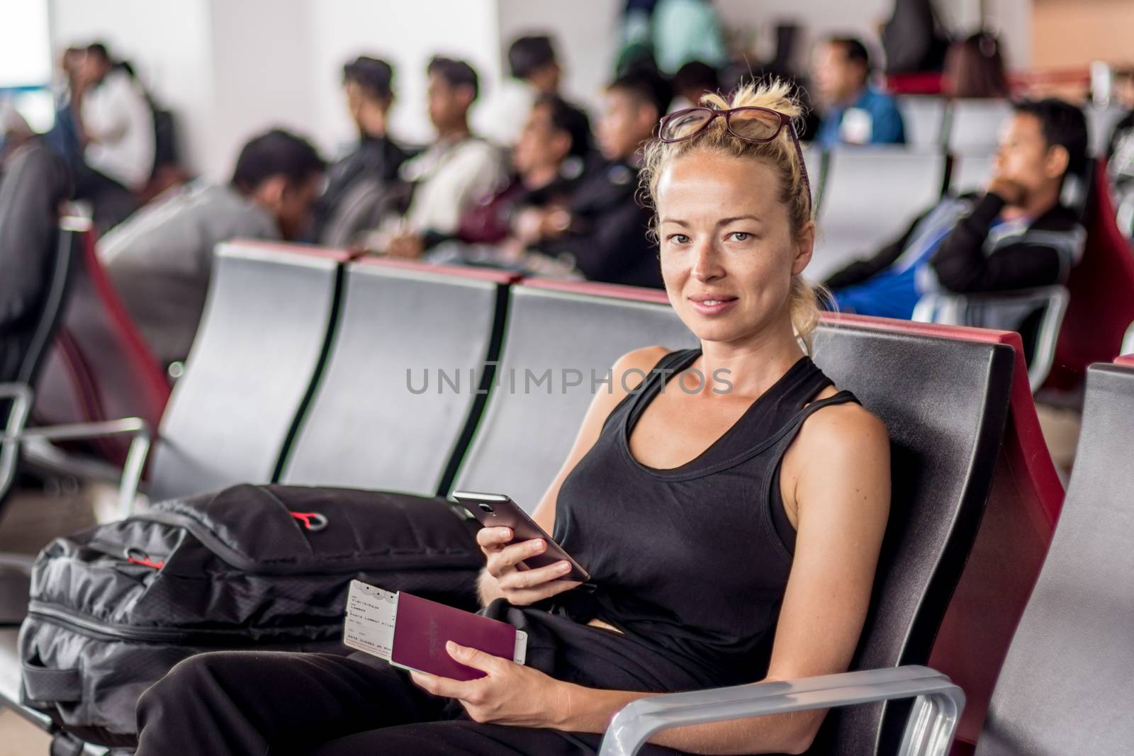Casual tanned blond female traveler holding cell phone, passport and boarding pass while waiting to board a plane at the departure gates at the asian airport terminal.