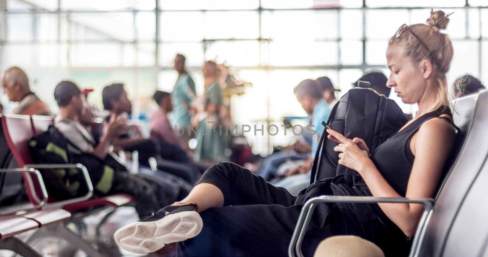 Casual sporty young blond female traveler using her cell phone while waiting to board a plane at the departure gates at the asian airport terminal.
