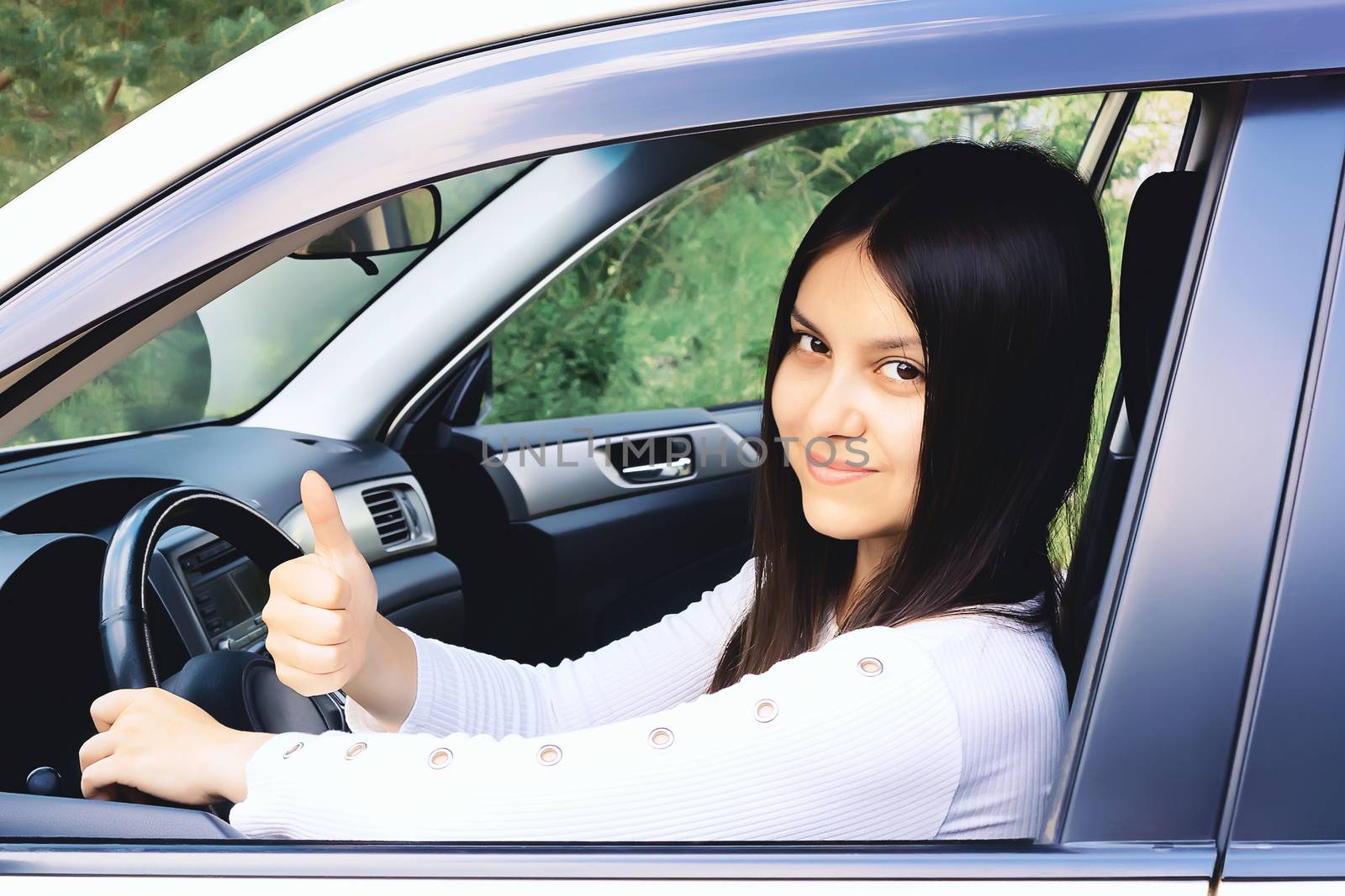 Happy young girl with long dark hair driving a car and gesturing thumb up by galsand