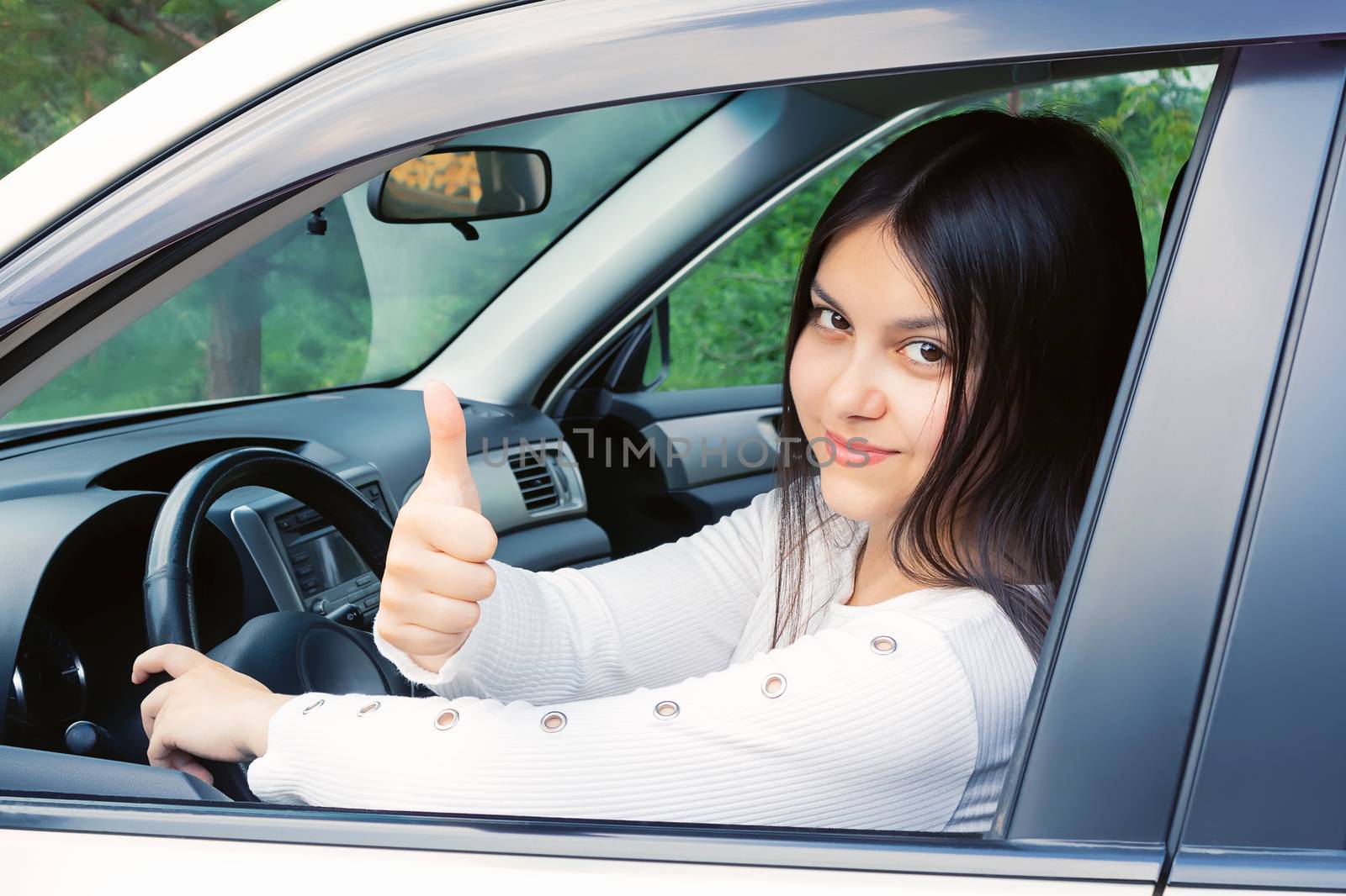 Happy young girl with long dark hair driving a car and gesturing thumb up.