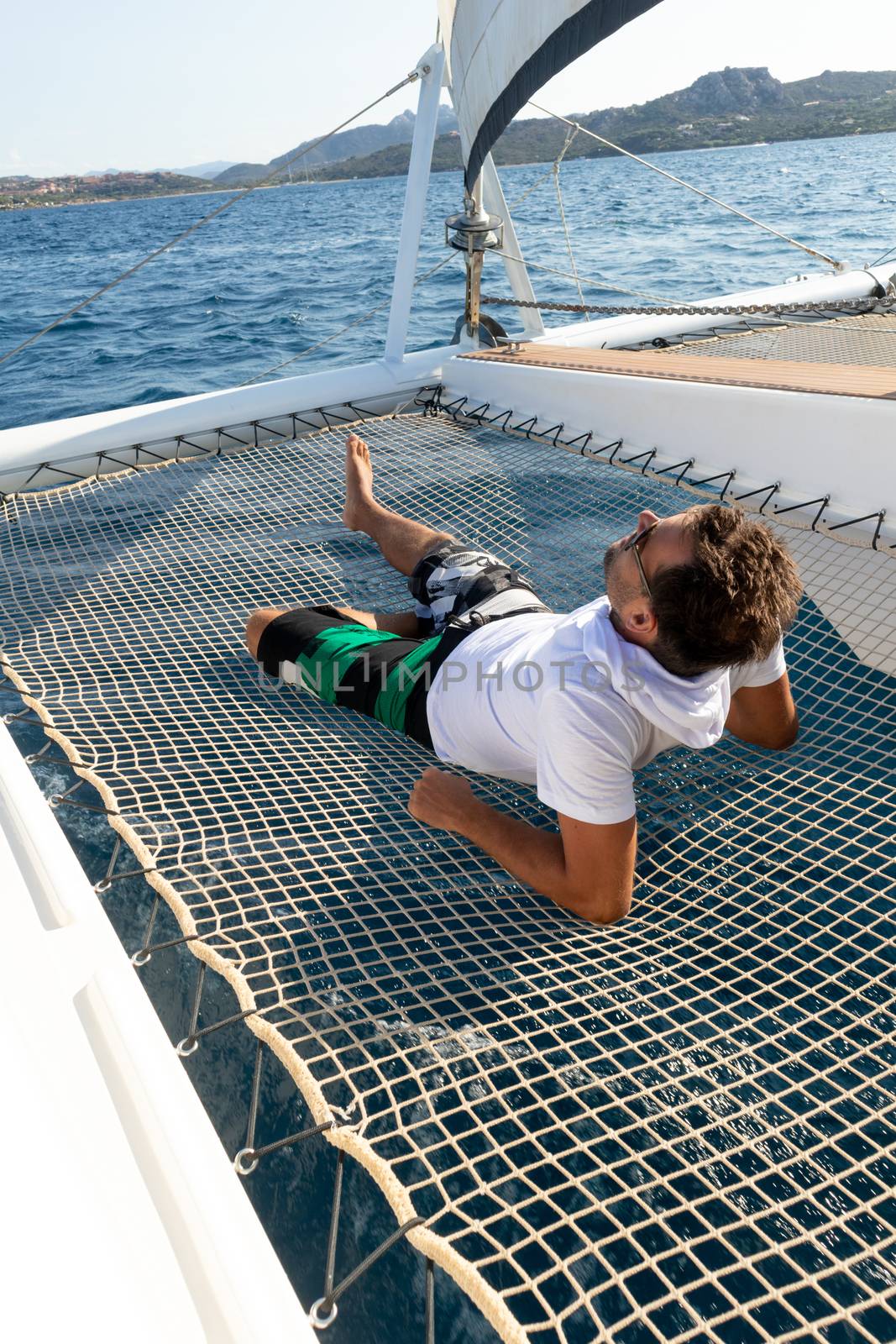 Sporty man relaxing, lying in hammock of a catamaran sailing boat on luxury nautic vacations near picture perfect Palau town, Sardinia, Italy.