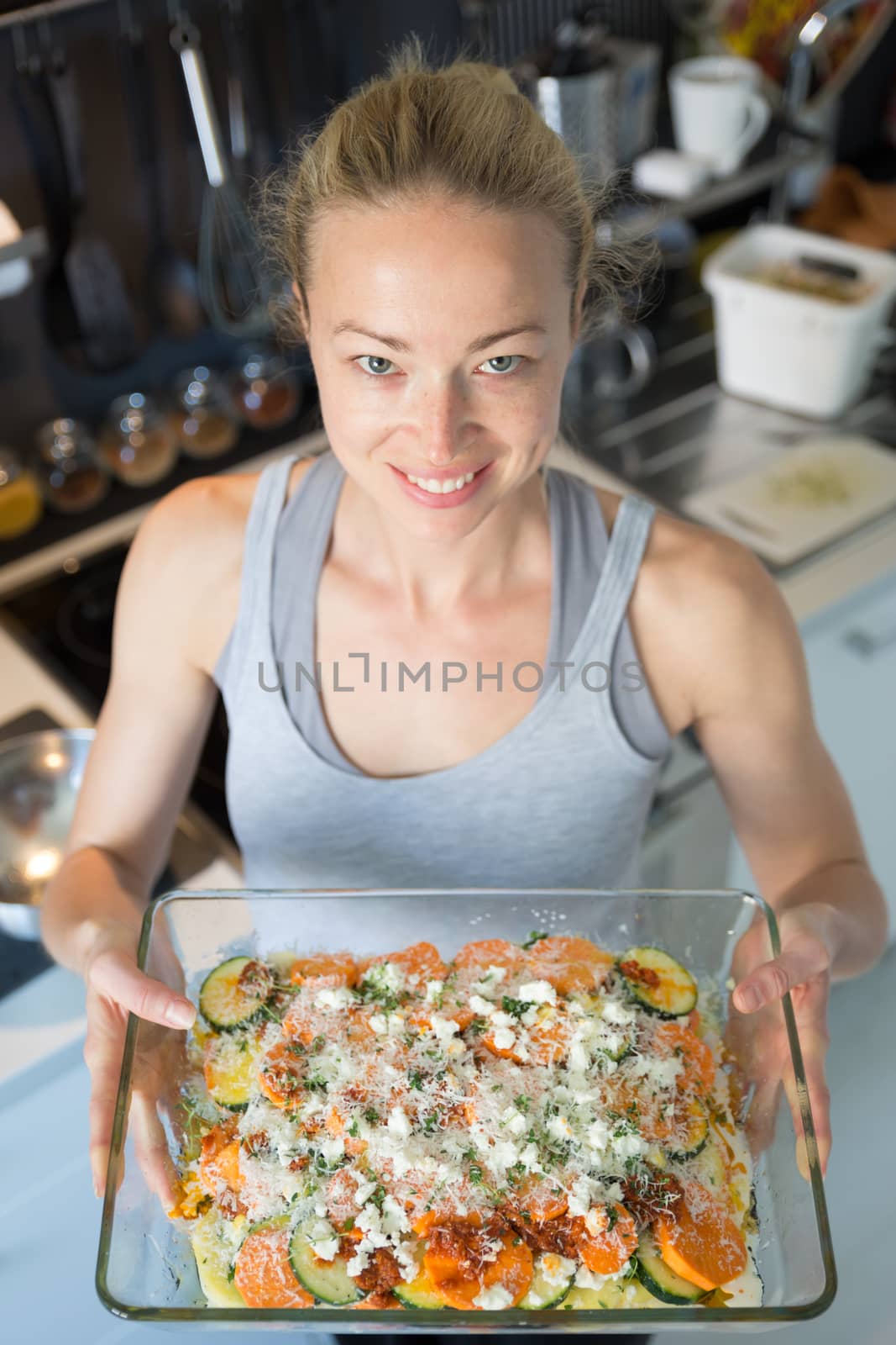 Smiling young healthy woman holding and proudly showing glass baking try with row vegetarian dish ingredients before placing it into oven. Healthy home-cooked everyday vegetarian food.