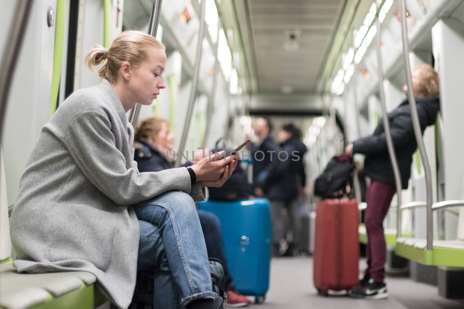 Beautiful blonde caucasian woman wearing winter coat reading on the phone while traveling by metro. Public transportation concept.