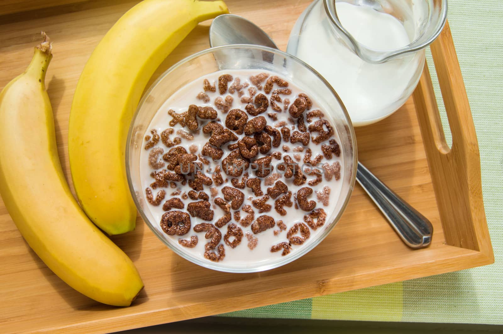 Bowl of oatmeal chocolate flakes in the shape of letters of the alphabet with milk on a wooden tray with bananas and a jug of milk by claire_lucia