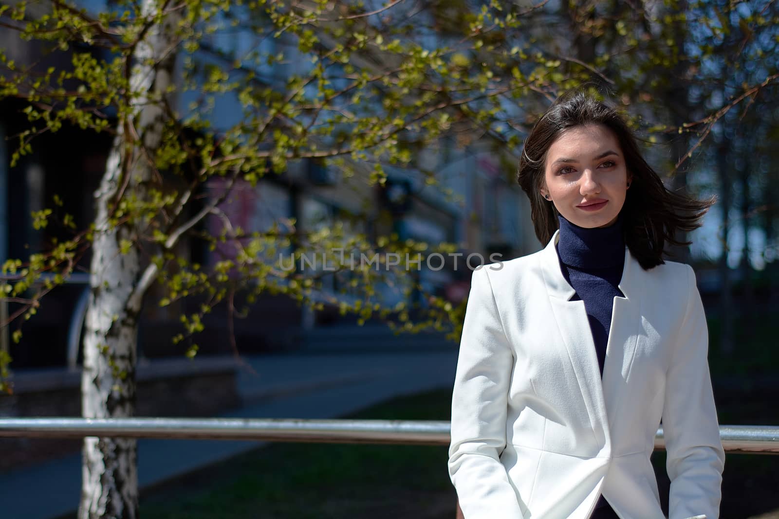 Elegant young woman dressed in a short white coat posing on a city street. Beautyful brunette woman. Modern urban woman portrait. Fashion business style clothes.