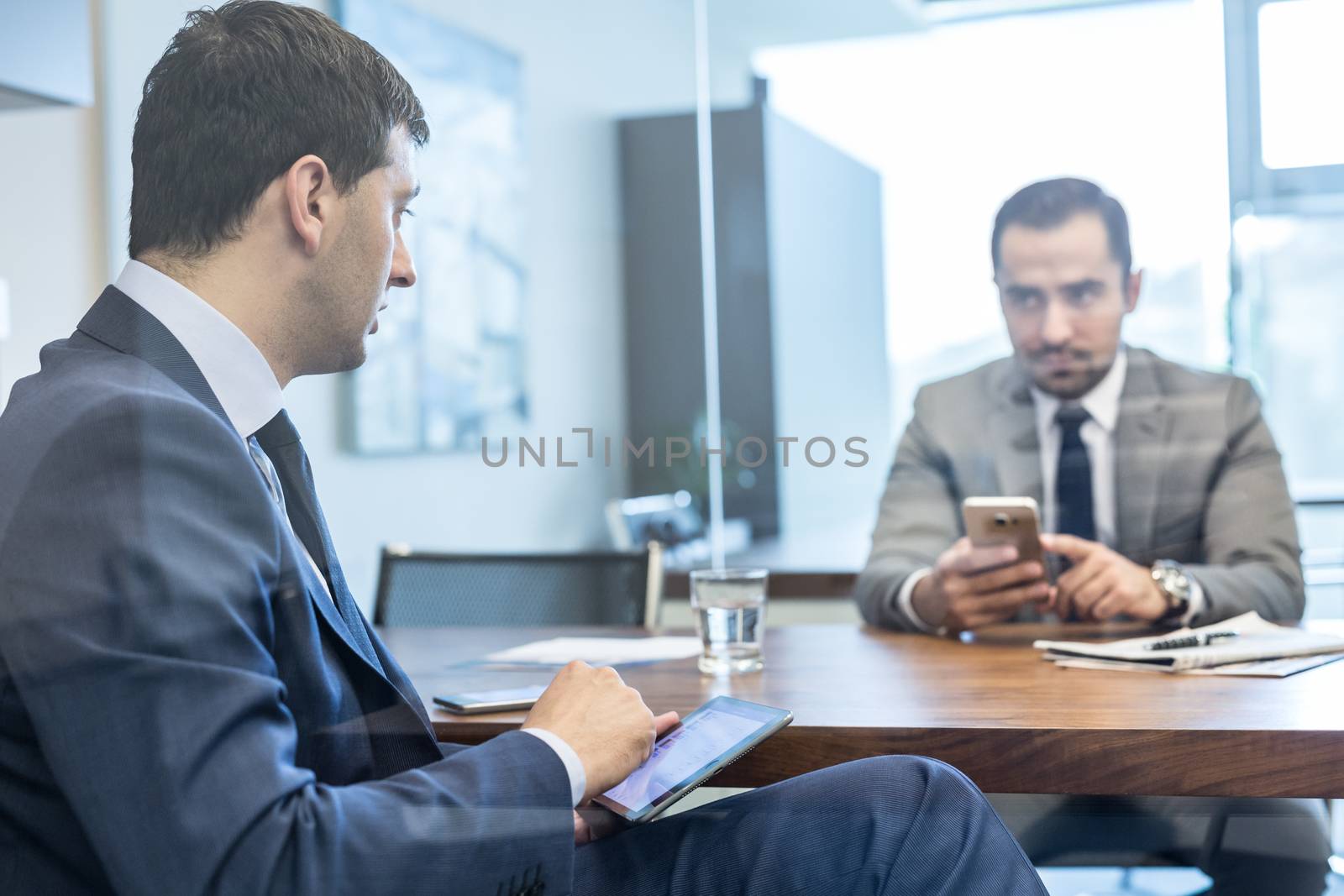 Through the window view of two young businessmen using tablet and smart phone devices at working business meeting in corporative office.